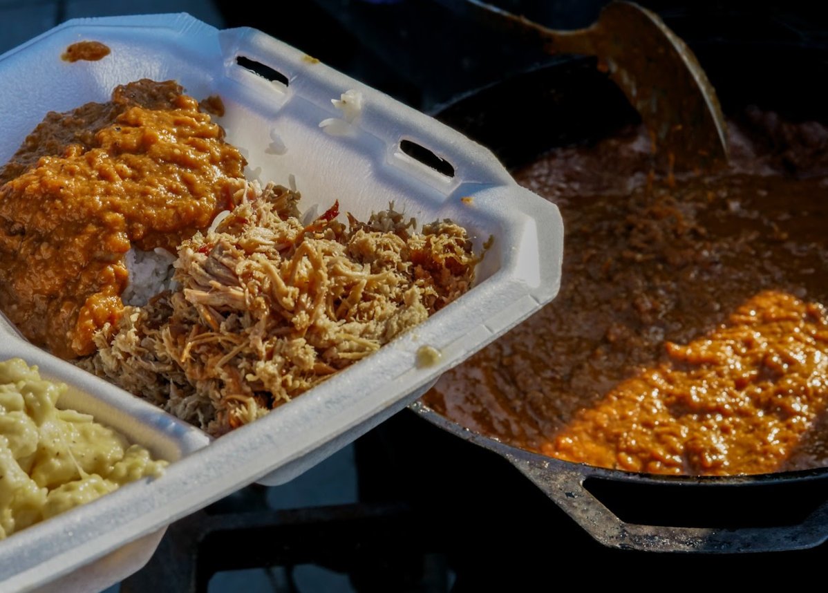 Takeout container with rice, shredded meat, gravy, and mashed potatoes next to a large pan with sauce.