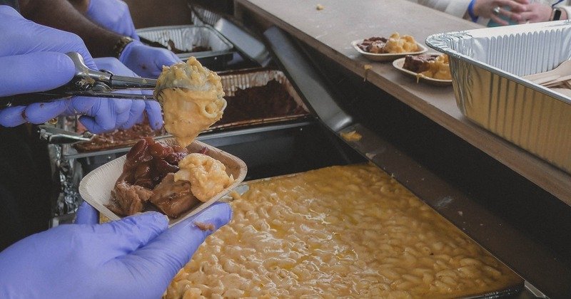 Person serving macaroni and cheese with barbecue meat into a small container, wearing blue gloves, at a buffet.
