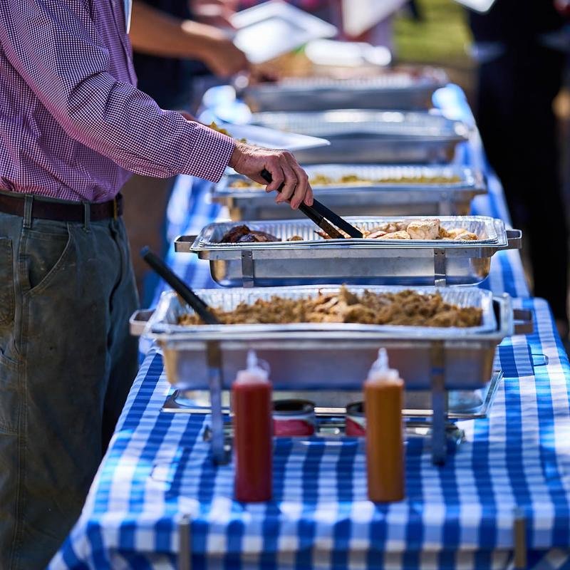 Person serving food from a buffet line with trays on a table covered with a blue and white checkered cloth.