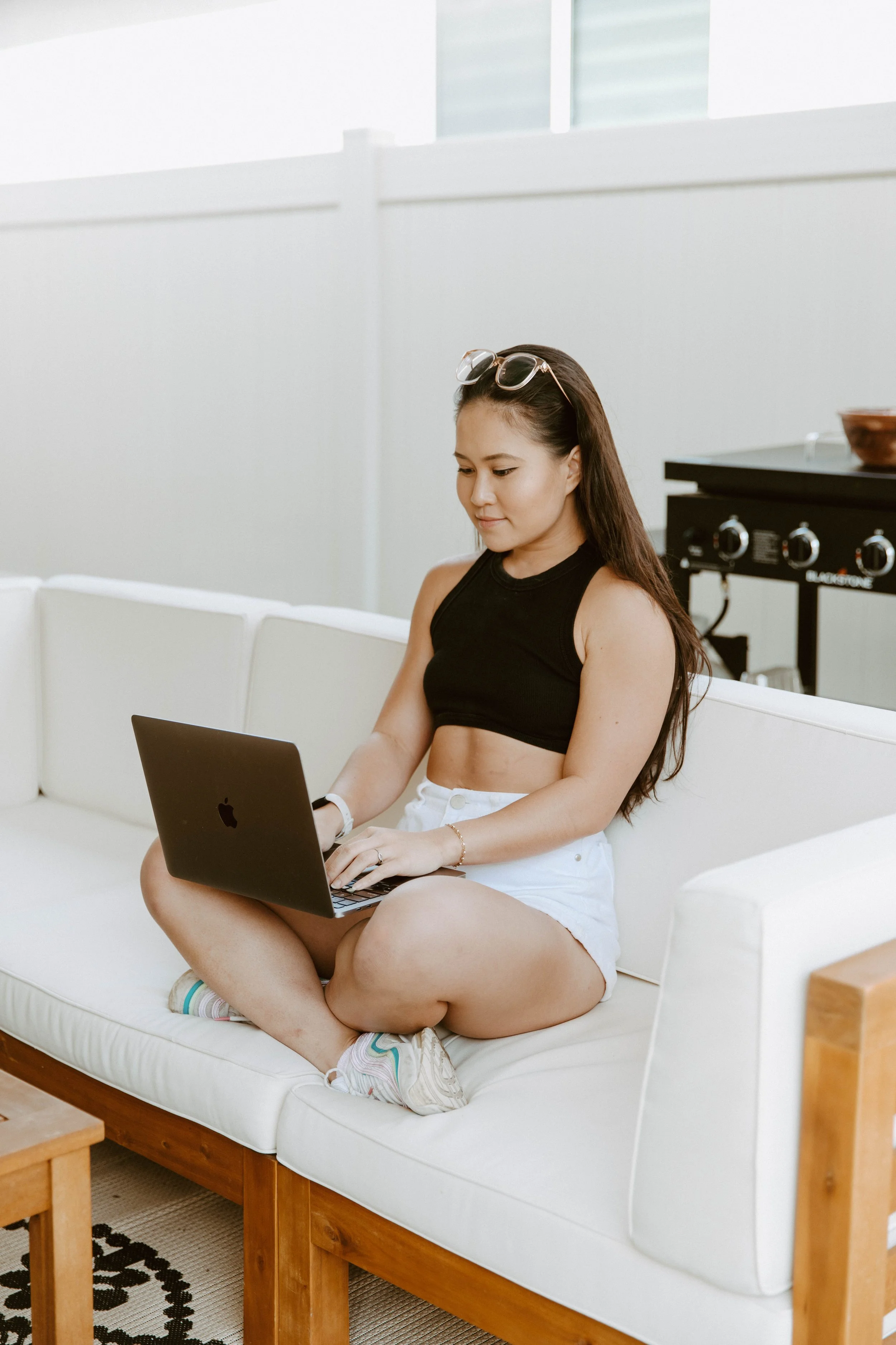 A young woman with long hair sitting cross-legged on a white couch using a black laptop. She is wearing a black crop top, white shorts, and white sneakers with colorful accents. Sunglasses are resting on her head, and she has a watch and a bracelet.