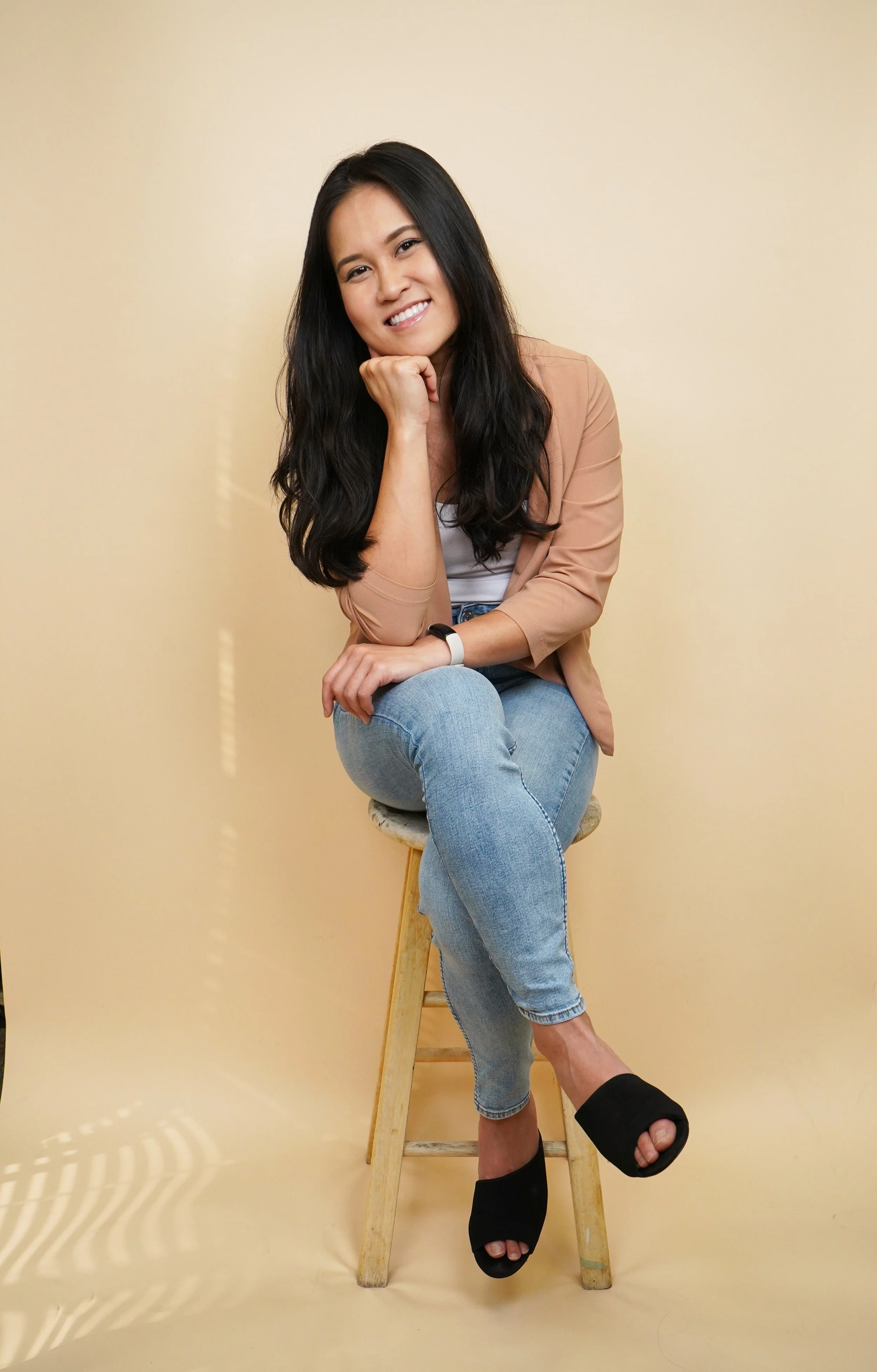 A woman with long black hair, smiling, sitting on a stool with her chin resting on her hand, wearing a beige blazer, white top, blue jeans, and black open-toe heels, against a plain beige wall.