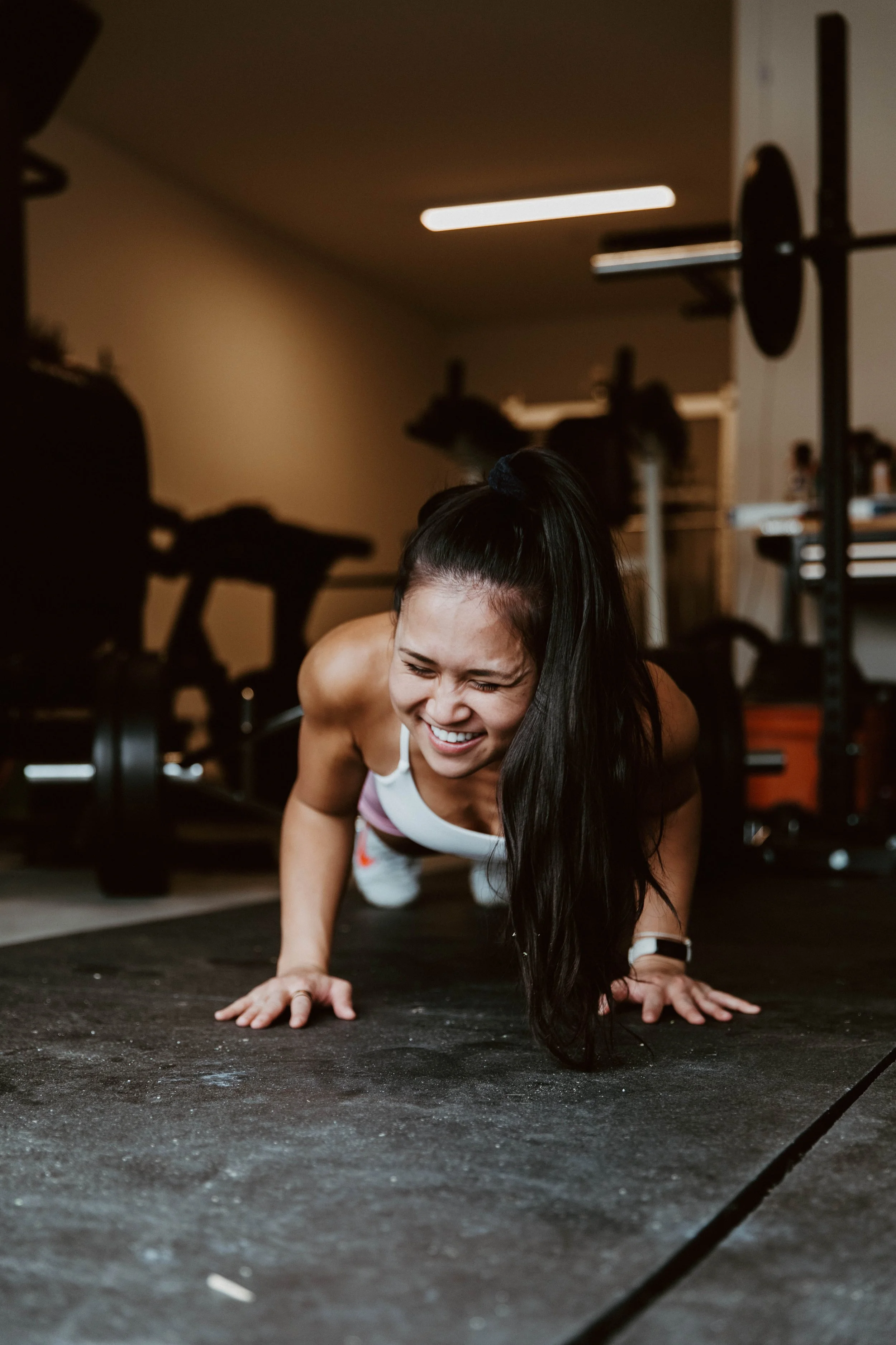Woman doing push-ups on gym floor, smiling, with gym equipment in background.