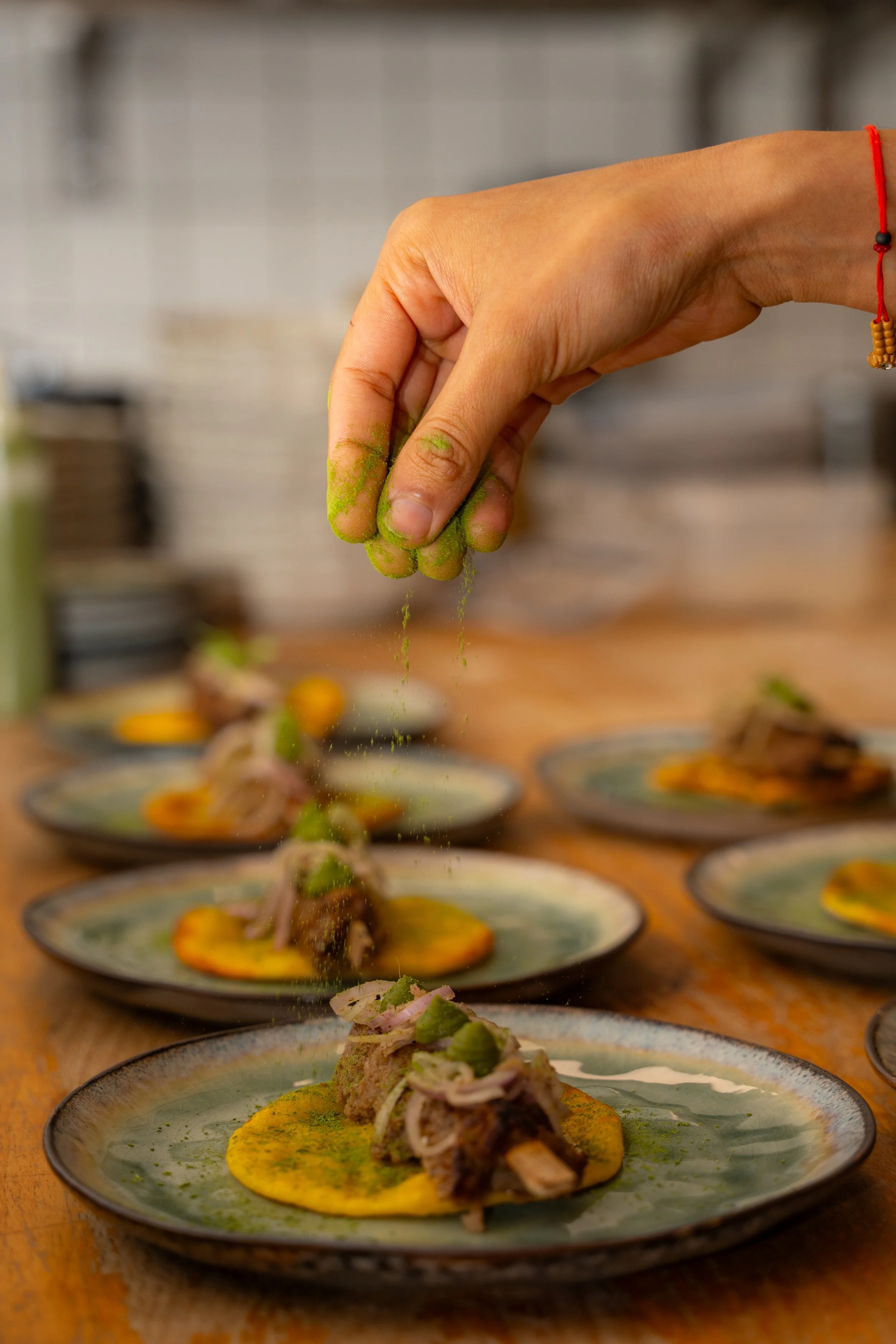 A hand sprinkling green powder onto a dish of food on a plate. Several similar plates are on the table.