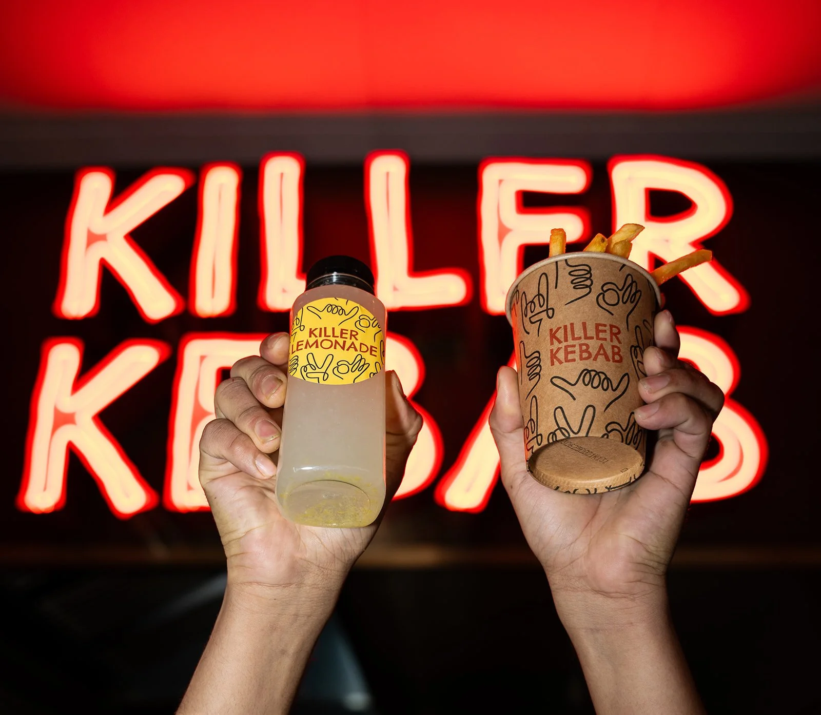 Lemonade og pommes frites holdes op foran det røde Killer Kebab-neonskilt