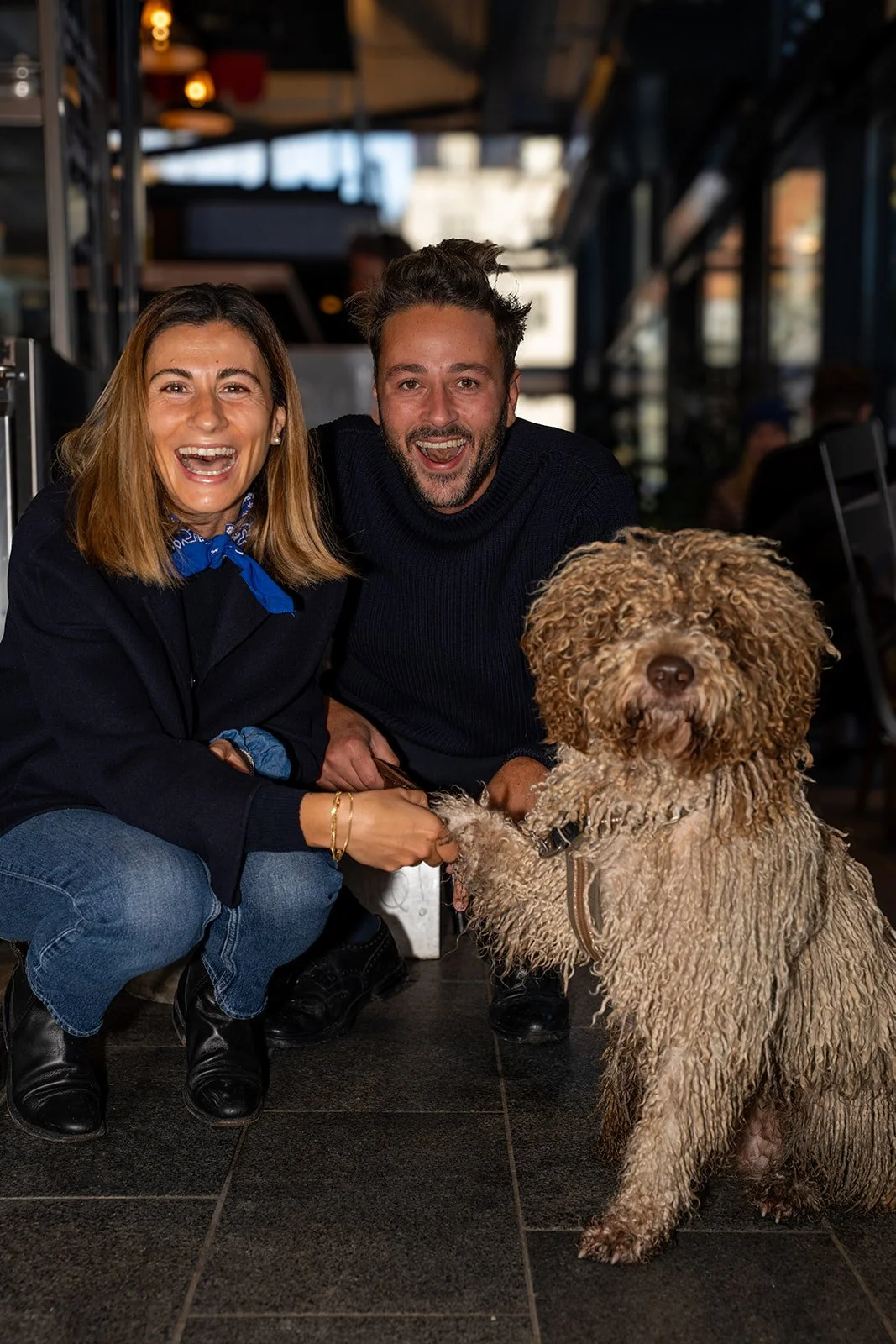 To smilende personer sidder på hug med en krøllet hund foran Il Mattarello
