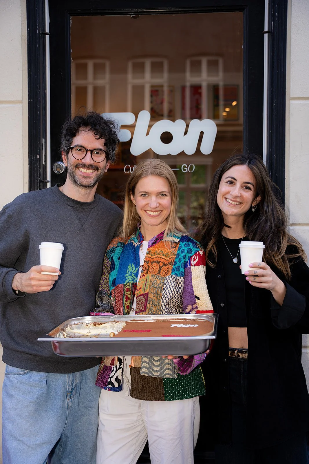 Tre smilende personer med kaffekopper og en bakke tiramisu foran caféen Flan