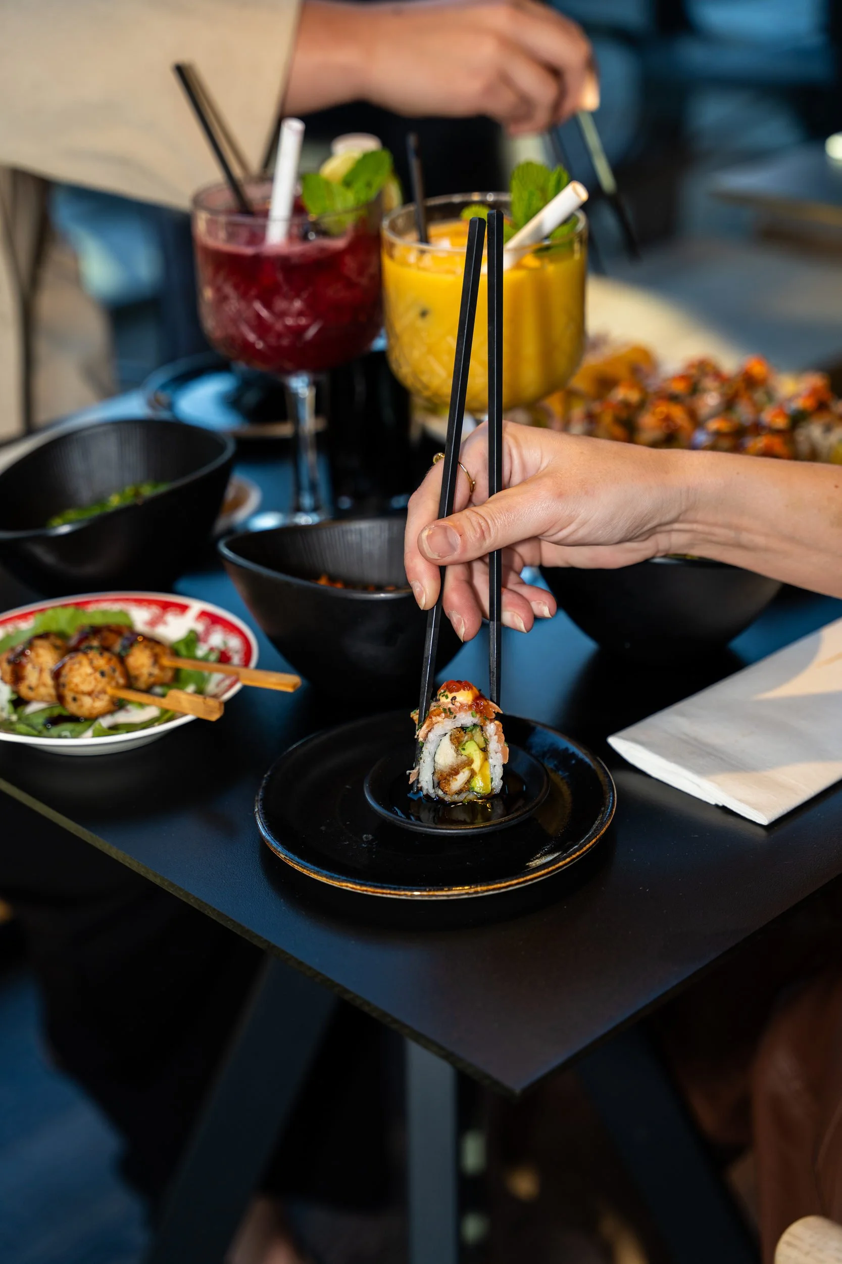 Spisepinde tager sushi fra fadet mellem røde og gule cocktails på Sushi Lab