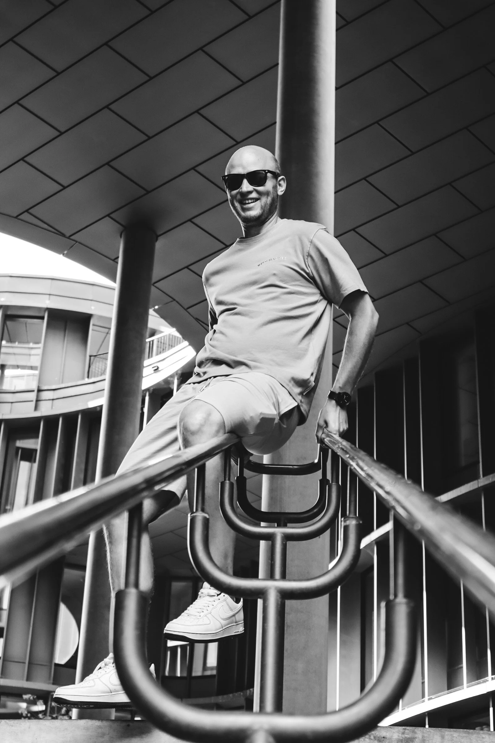 A smiling man in sunglasses sitting on a railing at an outdoor modern building with curved architectural design.