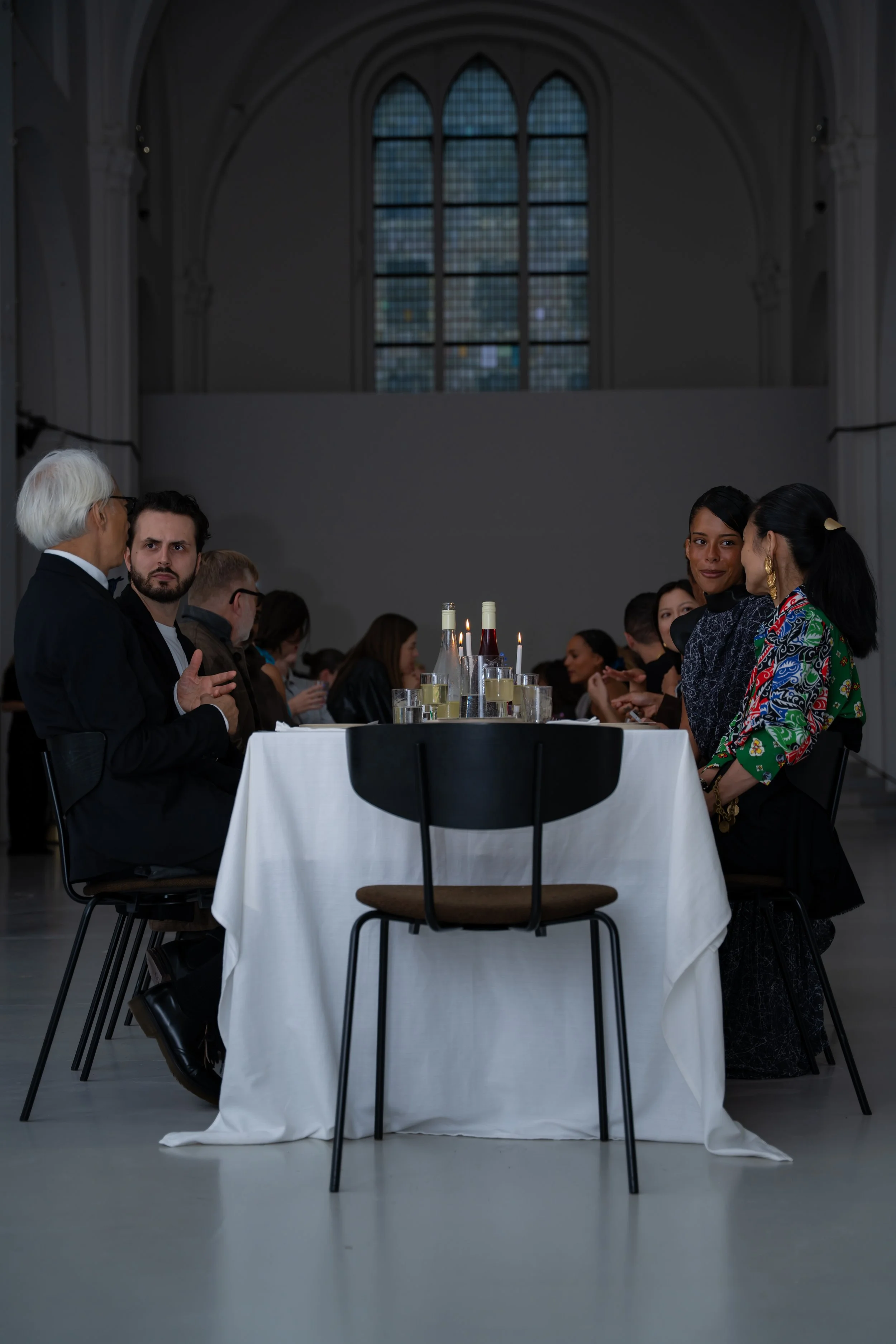 People seated at a banquet table with candles and bottles in a large hall with high arched windows.