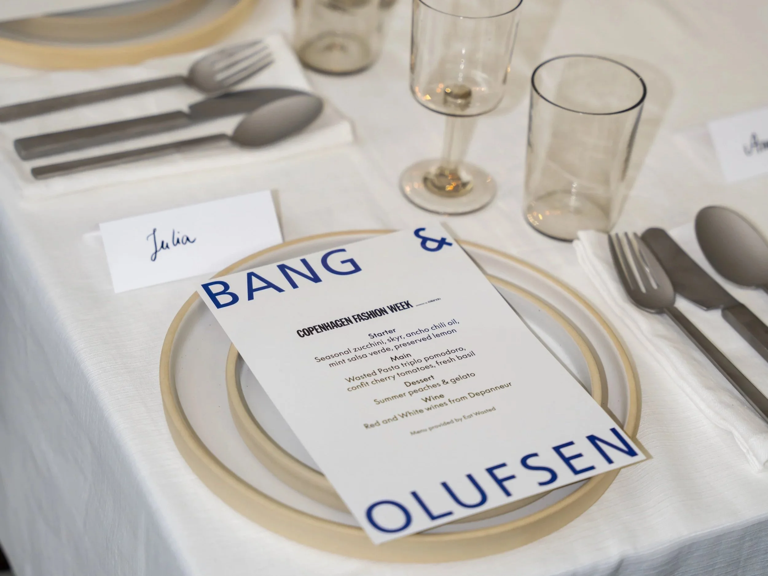 Table setting with a white tablecloth, a plate with a white and gold rim, a menu card, and silverware including a fork, knife, and spoon. Empty glasses and a place card with the name 'Julia' are also visible.
