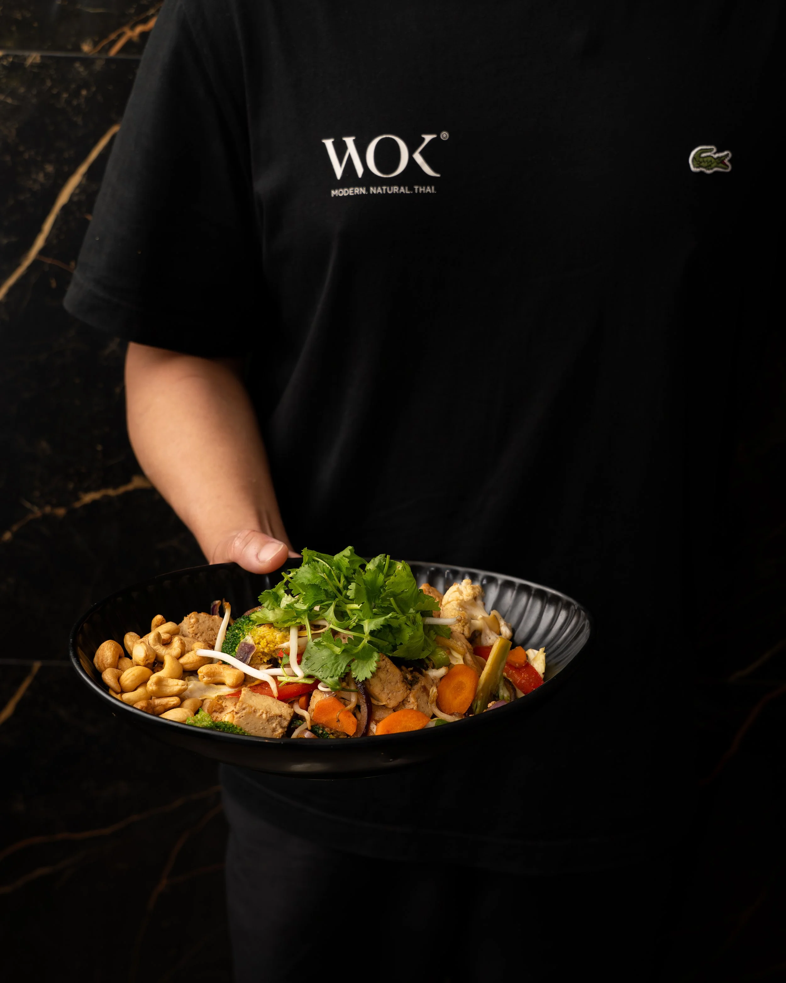 Person holding a black bowl of mixed vegetables and nuts, including carrots, lettuce, and sprouts, wearing a black shirt with the word 'WOK' and a small crocodile logo.