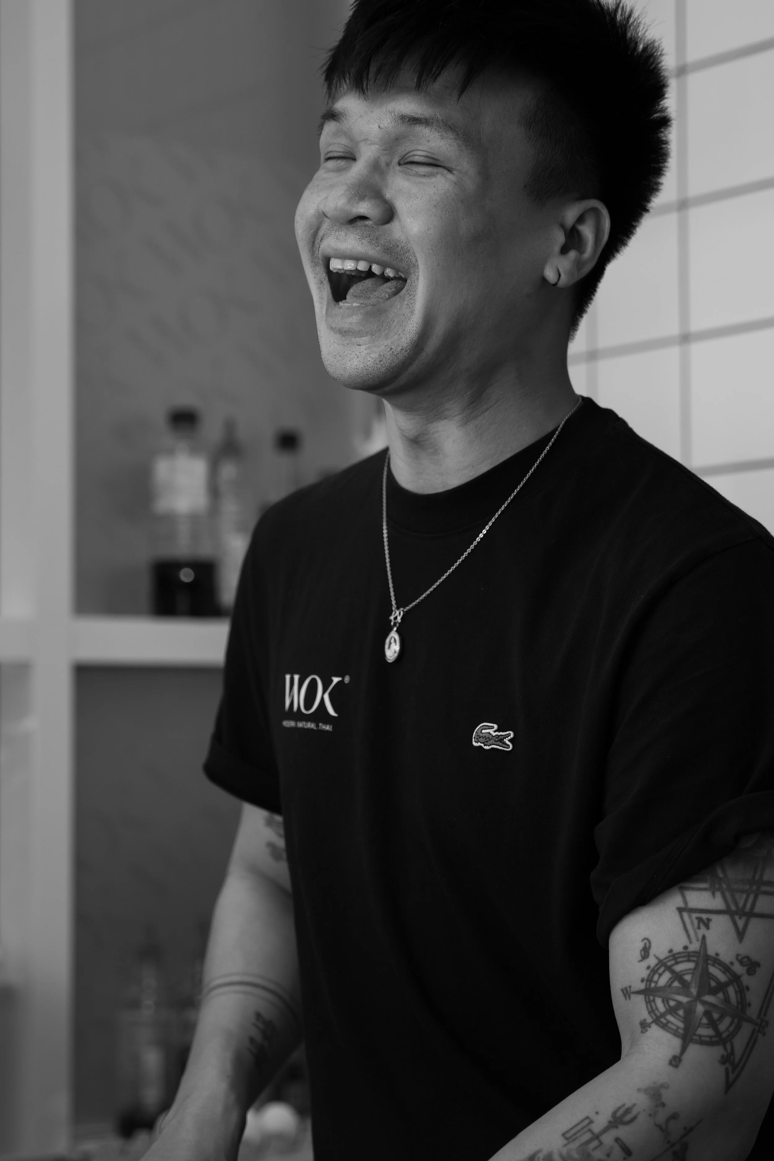 A man with short hairstyle laughing with eyes closed, wearing a black Lacoste t-shirt and a necklace, standing in a room with shelves and water bottles in the background.
