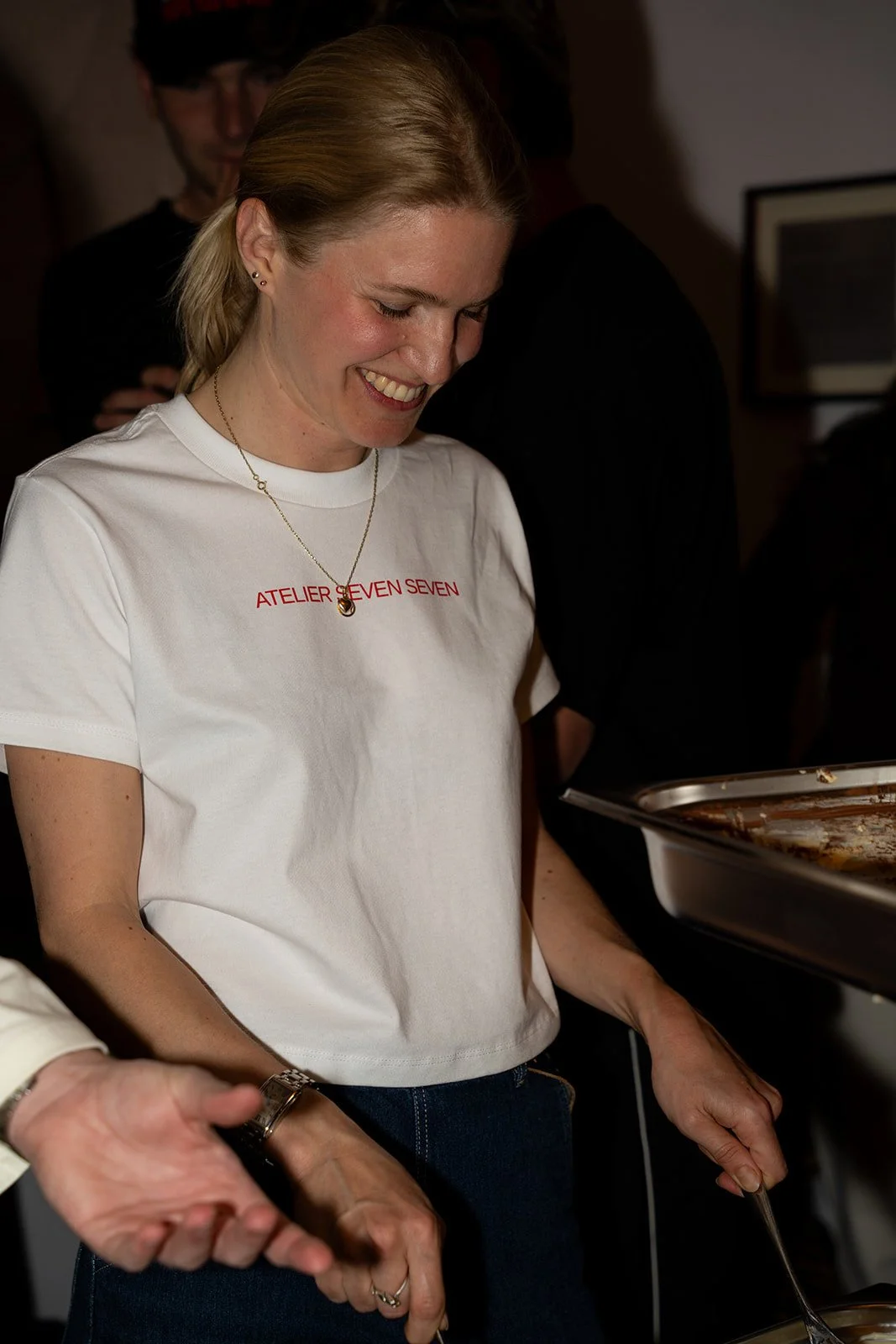 Smilende kvinde i hvid T-shirt serverer tiramisu fra bakken for gæster hos Arturel