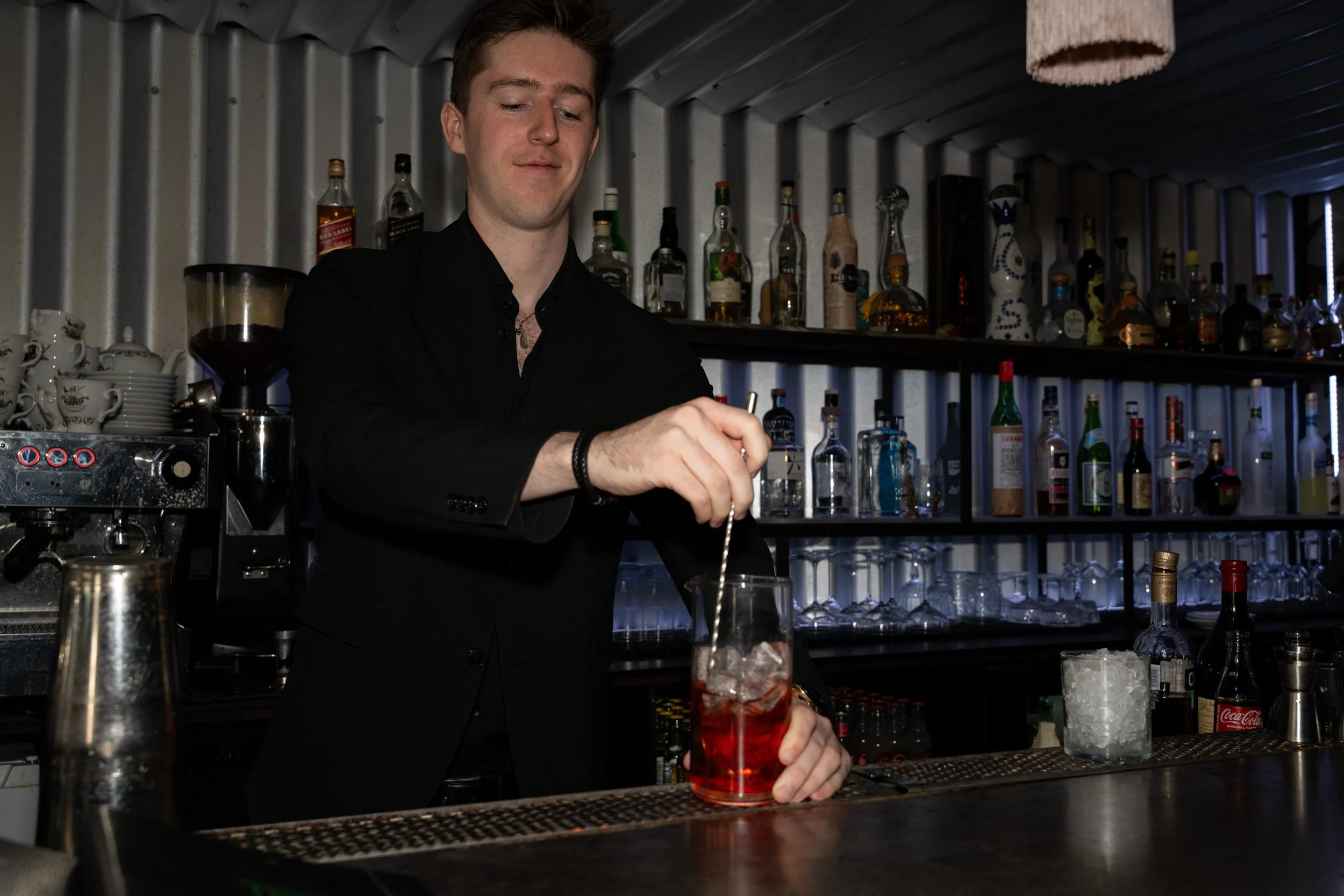 Bartender rører en rød cocktail foran de oplyste flaskehylder på Little Green Door København