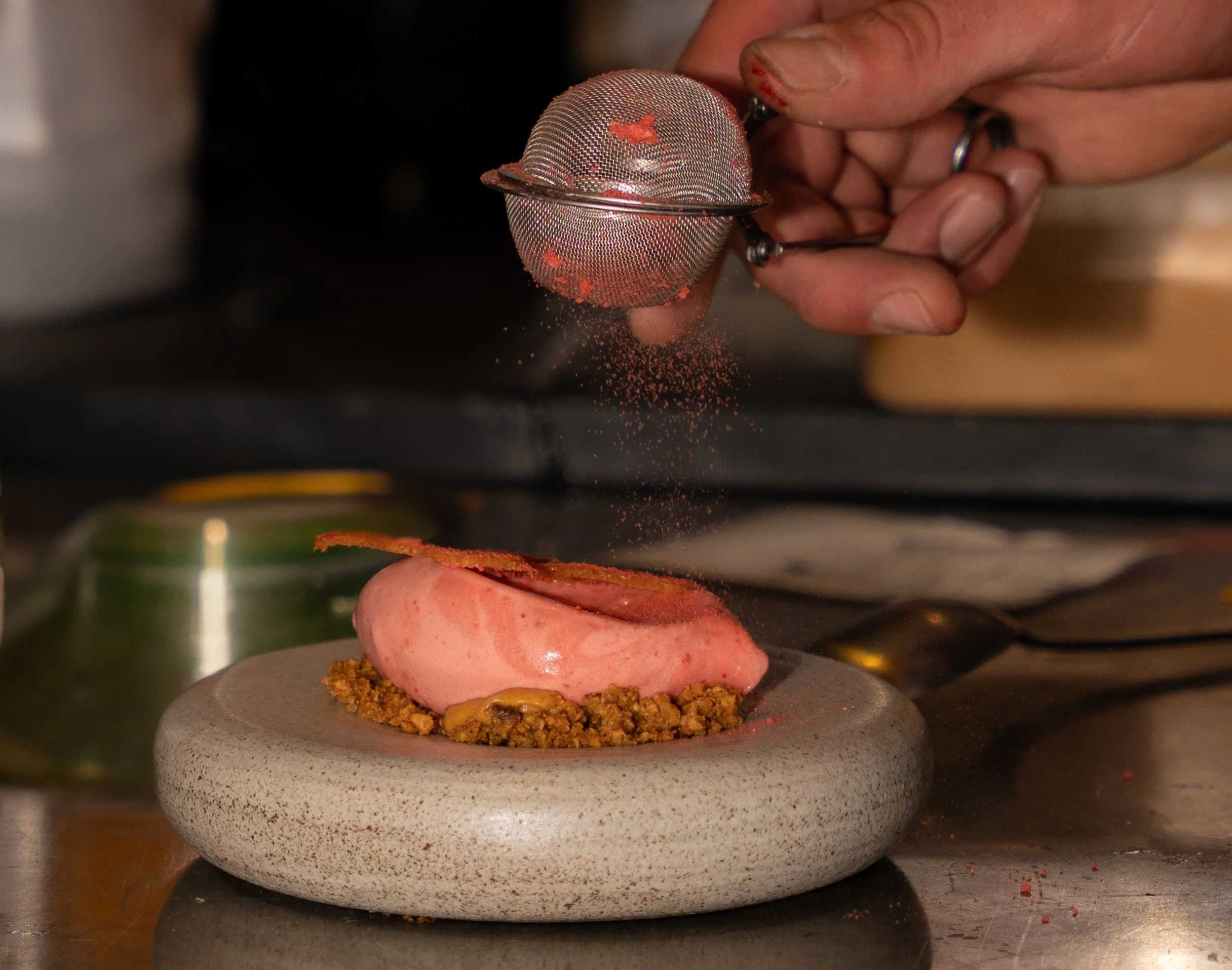 A chef sifts red powder onto a pink ice cream or mousse dessert garnished with a crisp, on a textured plate.