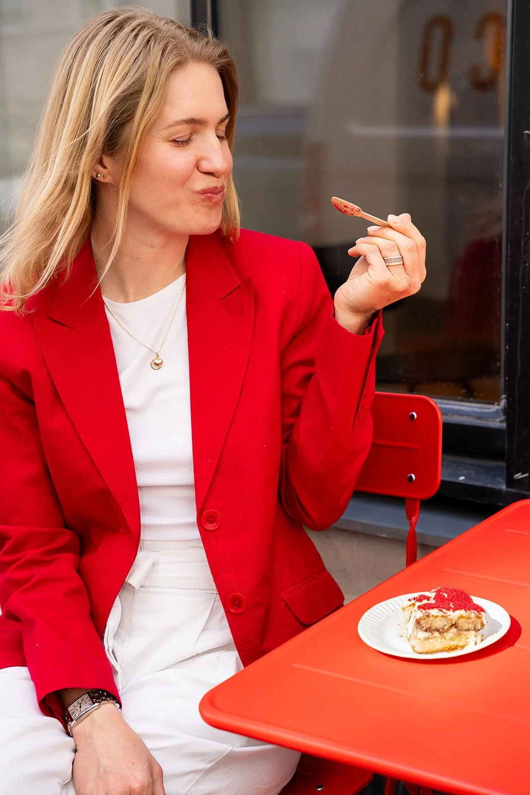 Kvinde i rød blazer smager på tiramisu ved et rødt bord foran caféen Flan
