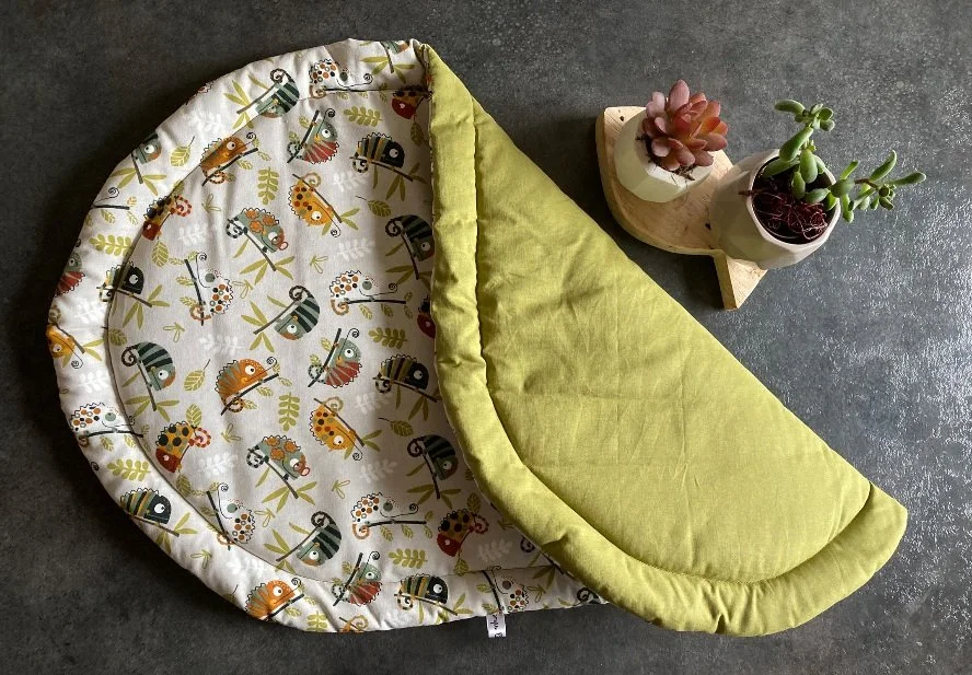 Un berceau ou lit pour bébé avec un motif d'animaux colorés et un côté vert, placé sur un sol en béton, à côté de deux petites plantes succulentes dans des pots blancs.