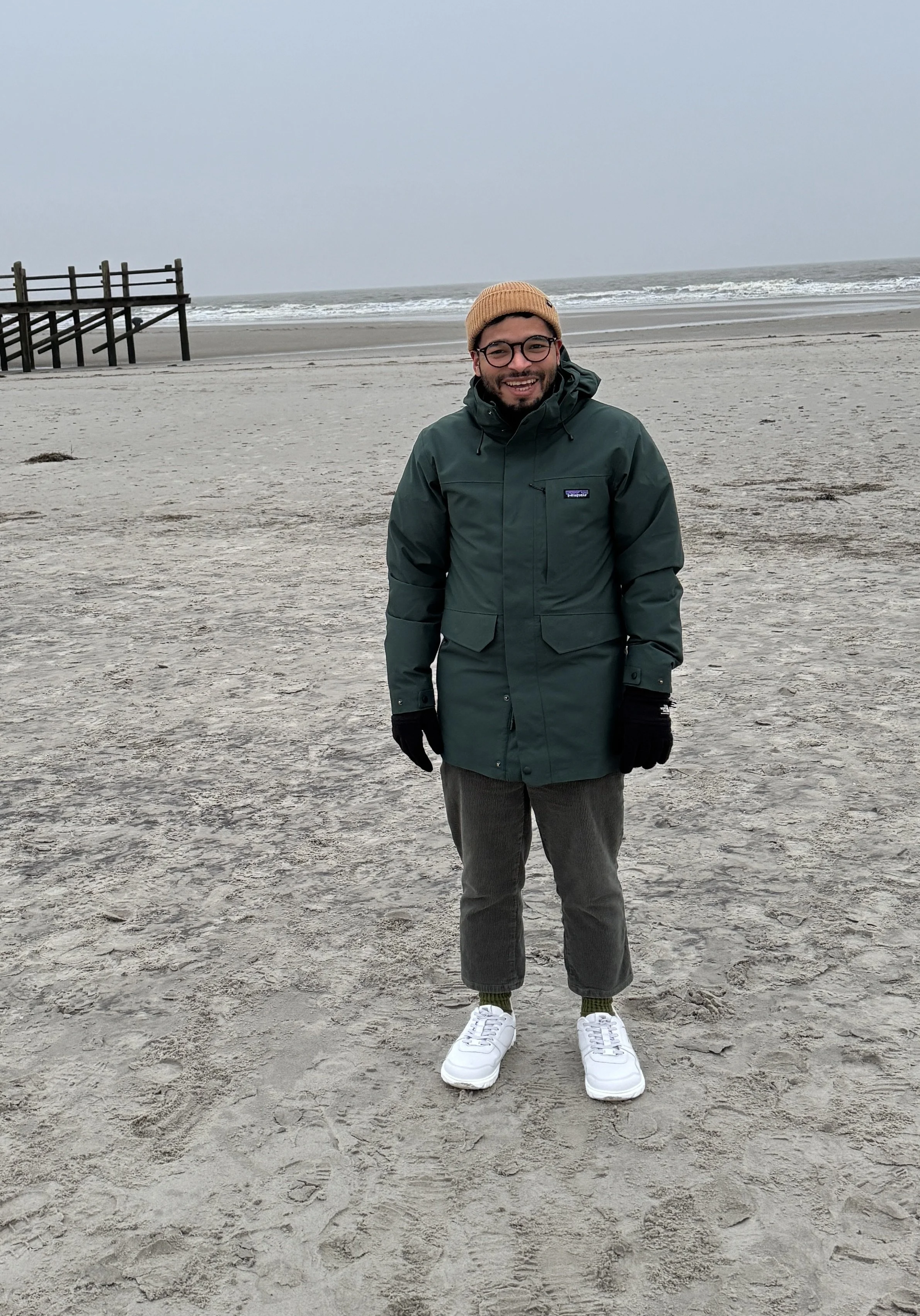 Mann auf einem Strand im Winter, trägt eine grüne Jacke, graue Hosen, schwarze Handschuhe, weiße Schuhe und eine braune Mütze, steht vor dem Meer.