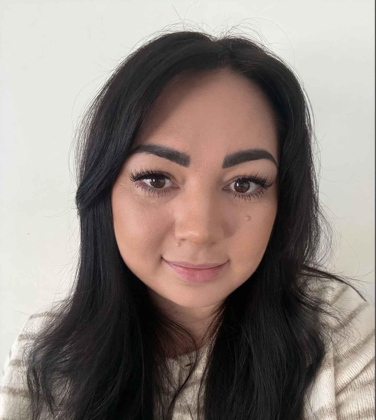 Close-up of a woman with long black hair, makeup, and a subtle smile, sitting near a window with blinds.