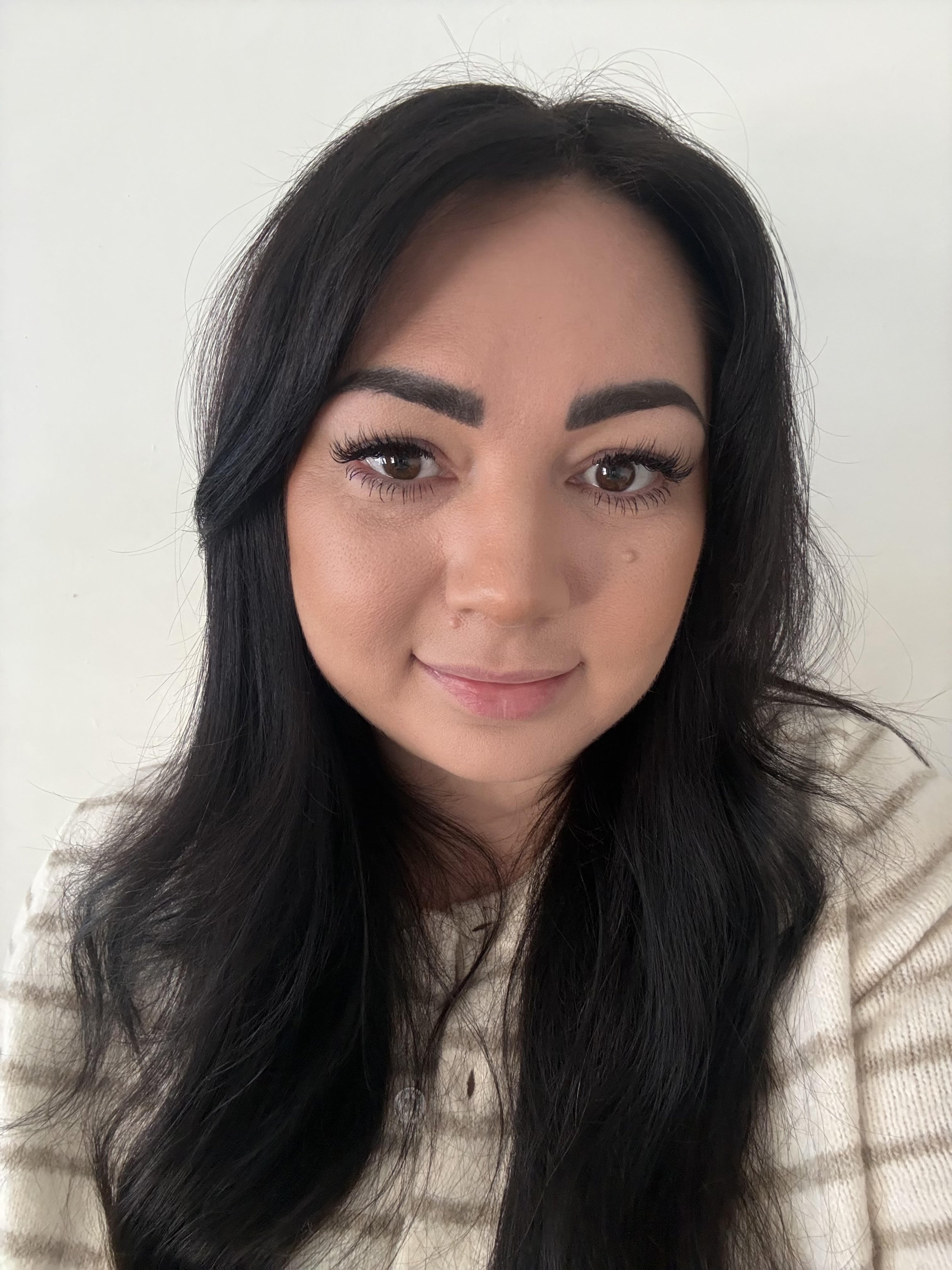 Close-up of a woman with long black hair, light skin, wearing makeup, smiling slightly, with a plain white background.