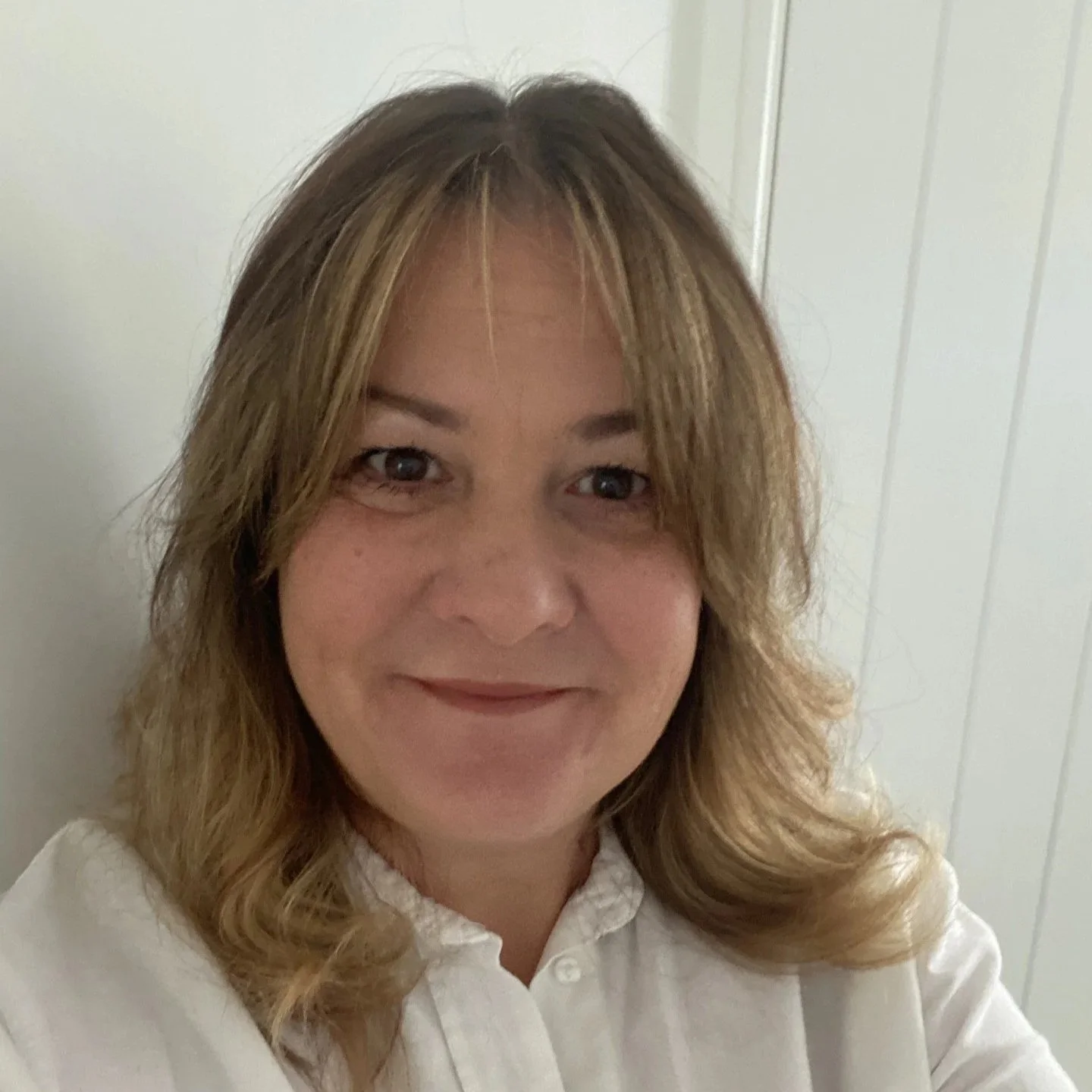 A smiling woman with shoulder-length light brown hair wearing a white collared shirt standing in front of a white-paneled wall.