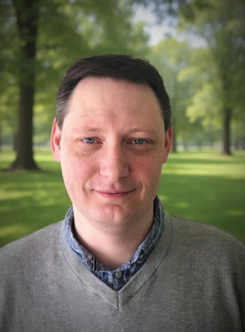 A man with short dark hair and blue eyes standing outdoors in a park with green trees and grass in the background.