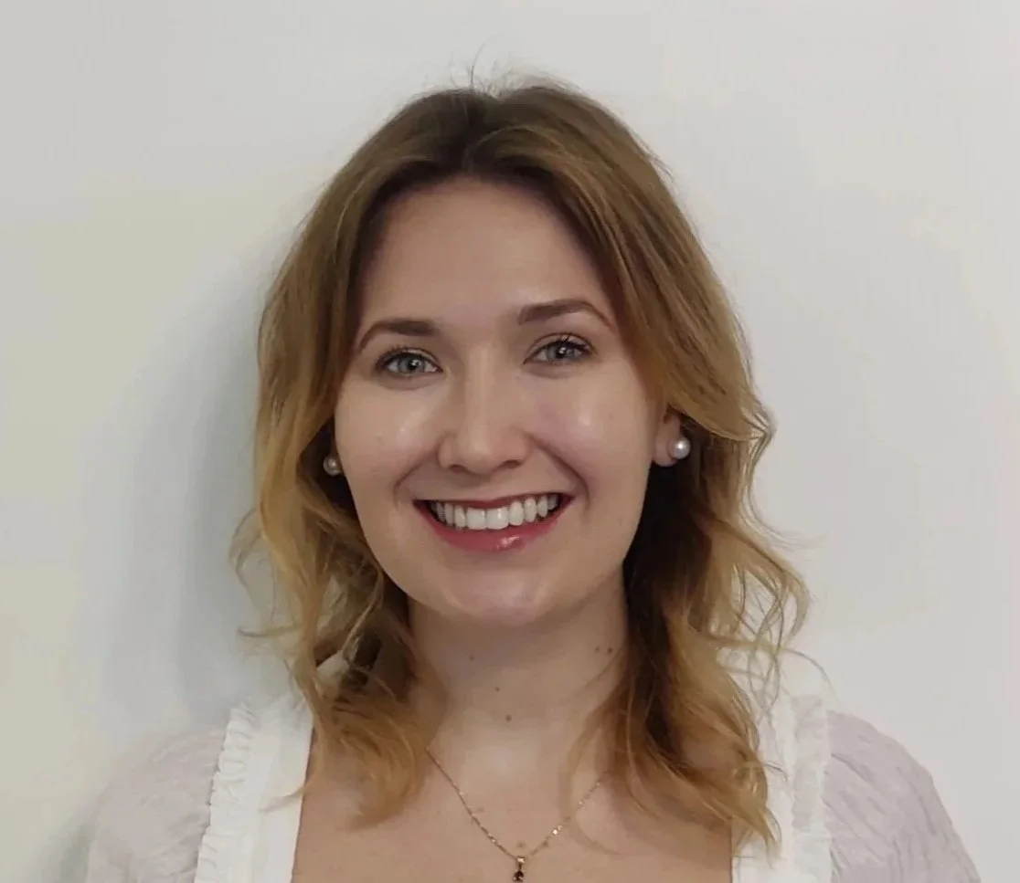 A smiling woman with shoulder-length wavy blonde hair, wearing pearl earrings and a delicate necklace, standing against a plain light-colored background.