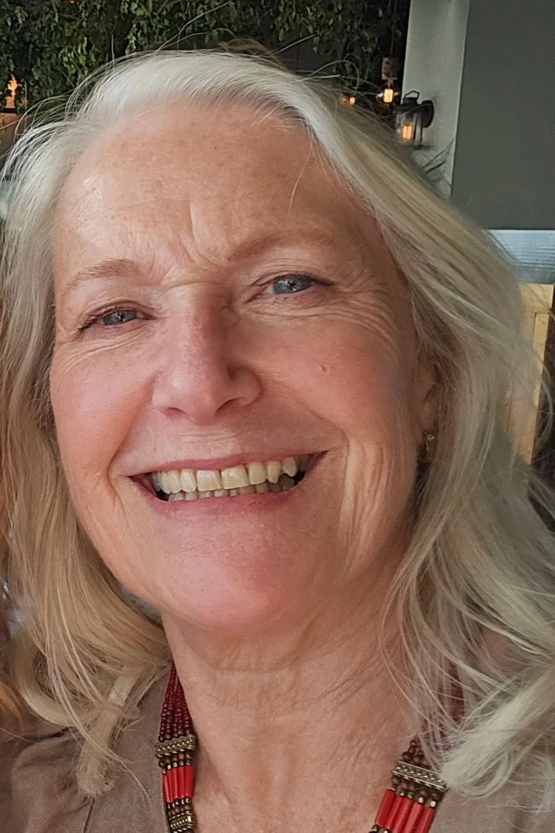 Close-up of an older woman with blonde hair, smiling with visible teeth, in a cheerful setting.