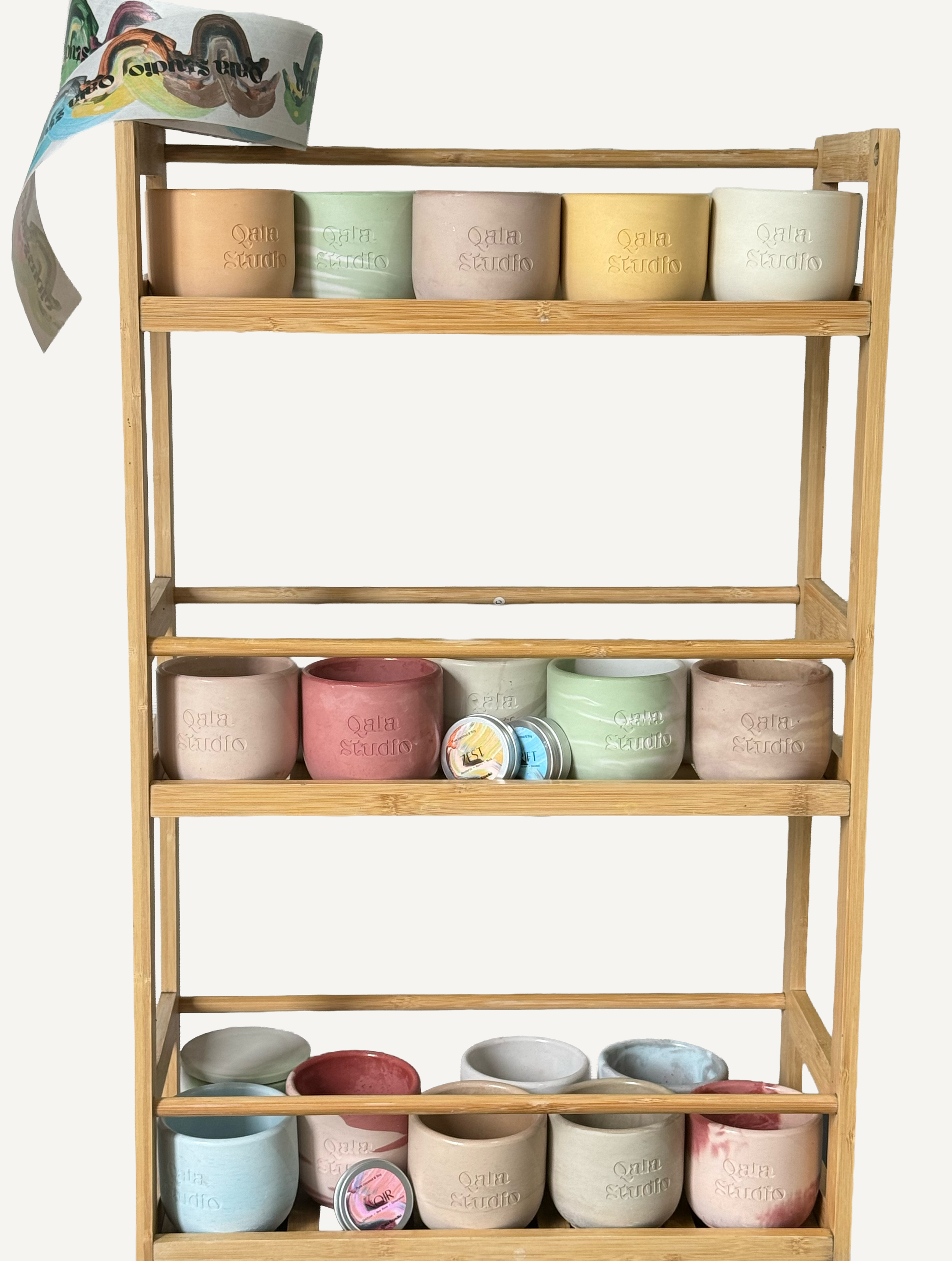 A wooden shelf holding pastel-colored ceramic bowls with 'Qala Studio' embossed, some with lids, arranged on multiple levels with a piece of colorful paper or fabric draped on the top left corner.