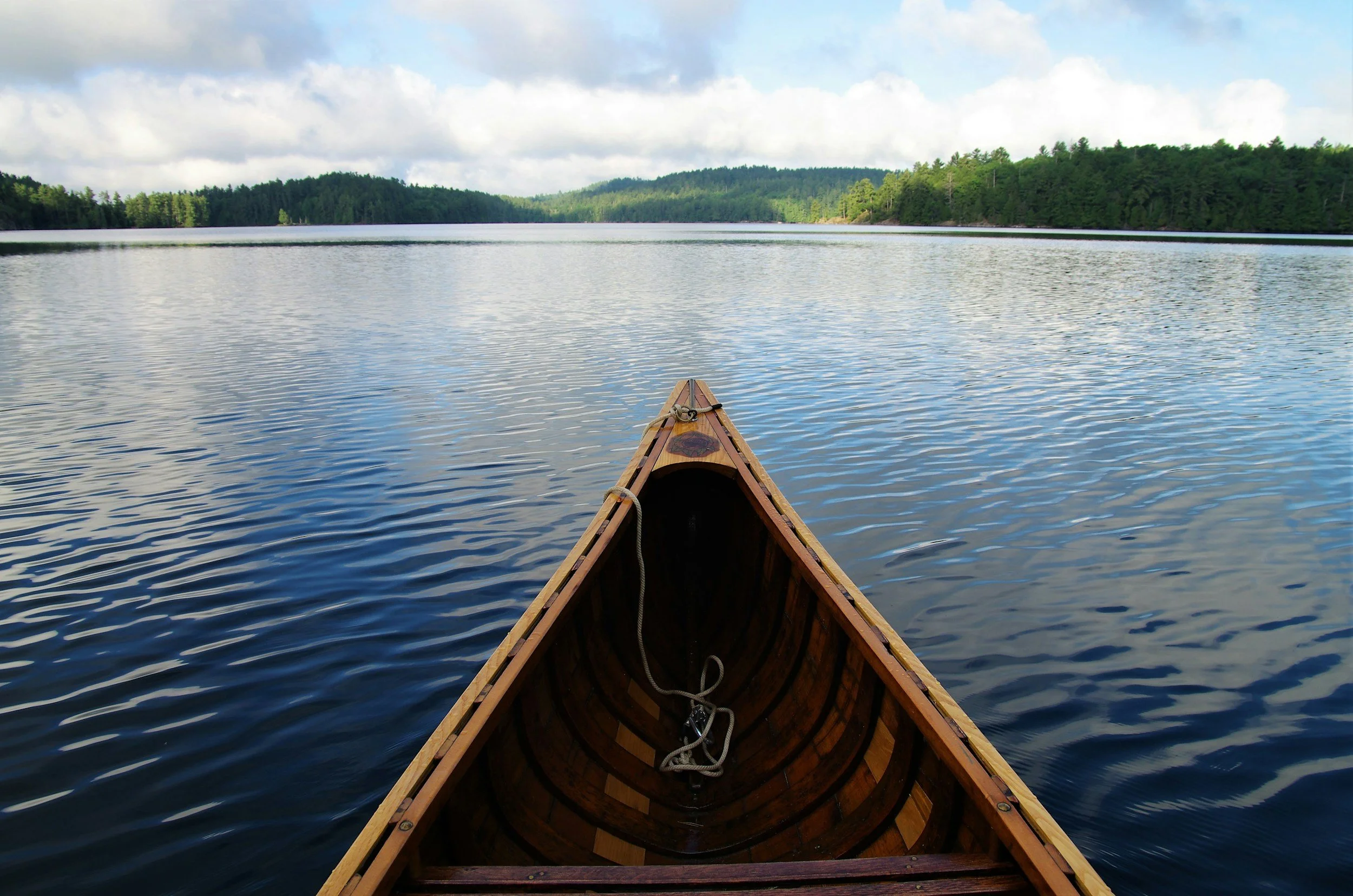 View from the front of a canoe on a calm lake, with green trees along the shoreline and partly cloudy sky.