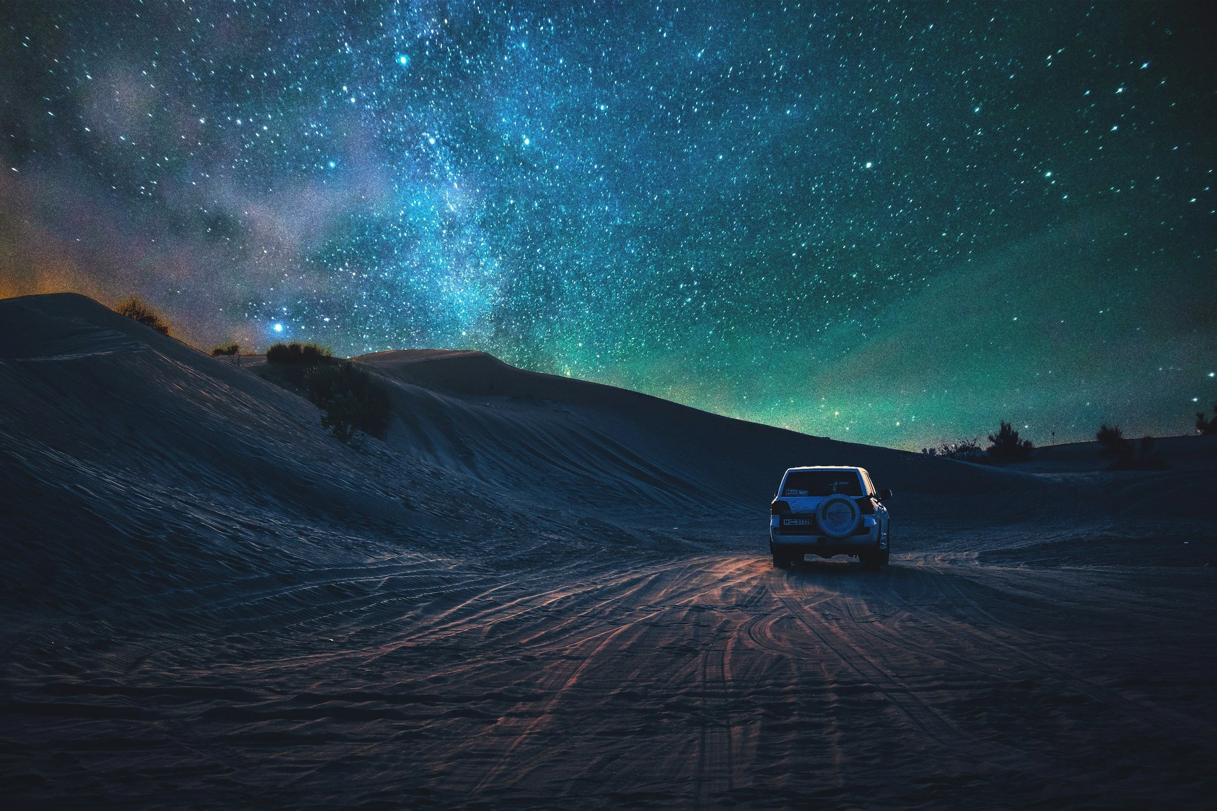 A vehicle driving on a sandy trail through sand dunes under a night sky filled with stars and the Milky Way.