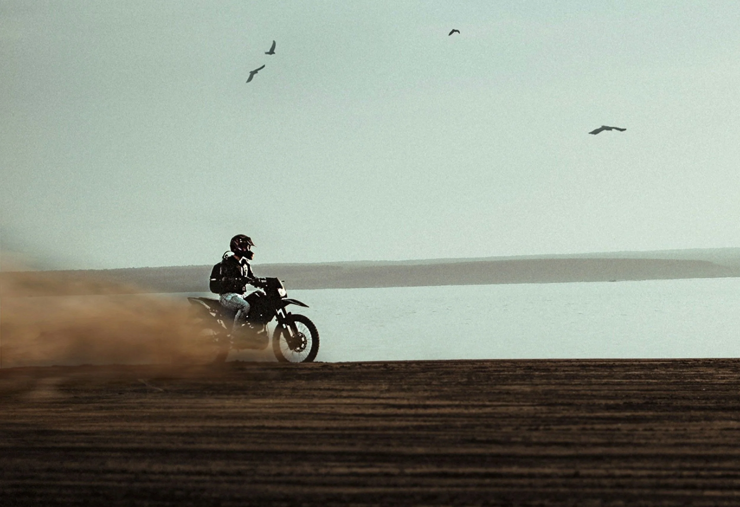 A person riding a dirt bike on a dirt trail by a body of water with birds flying in the sky.