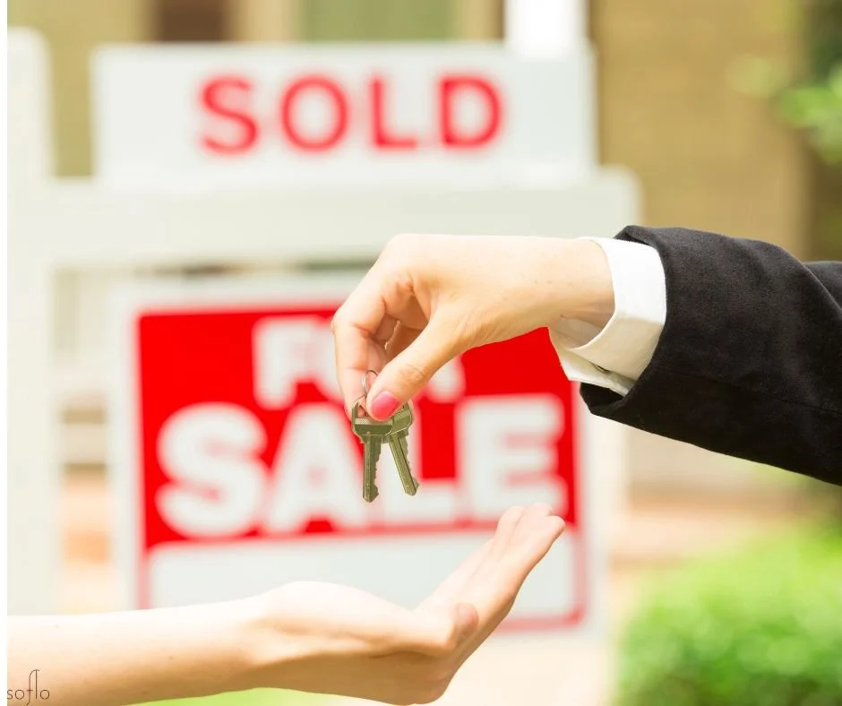 Person handing over keys to another person in front of a 'For Sale' sign with a 'Sold' sign in the background.