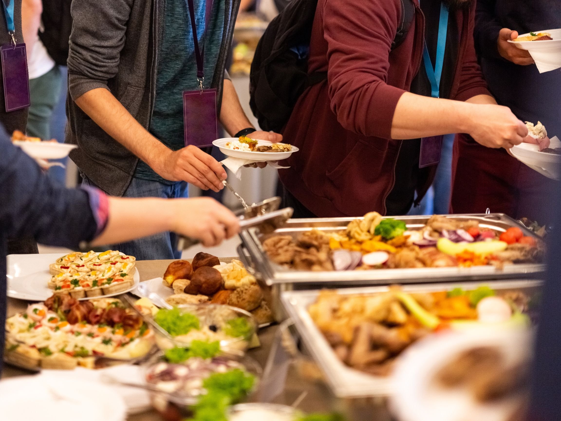 People serving themselves food at a buffet with various dishes including pizza, roasted meats, and vegetables.