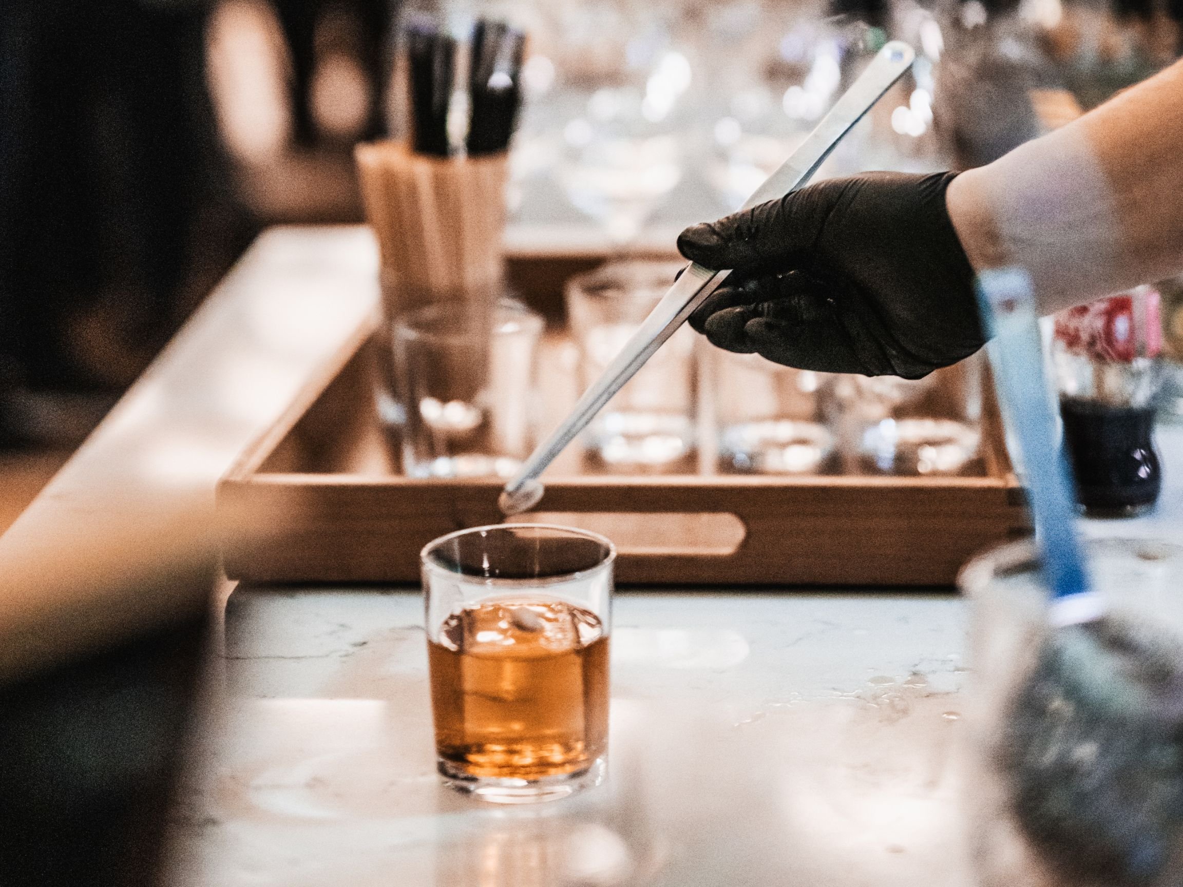 Person wearing black glove holding a metal tongs, serving a drink in a glass on a bar or countertop.