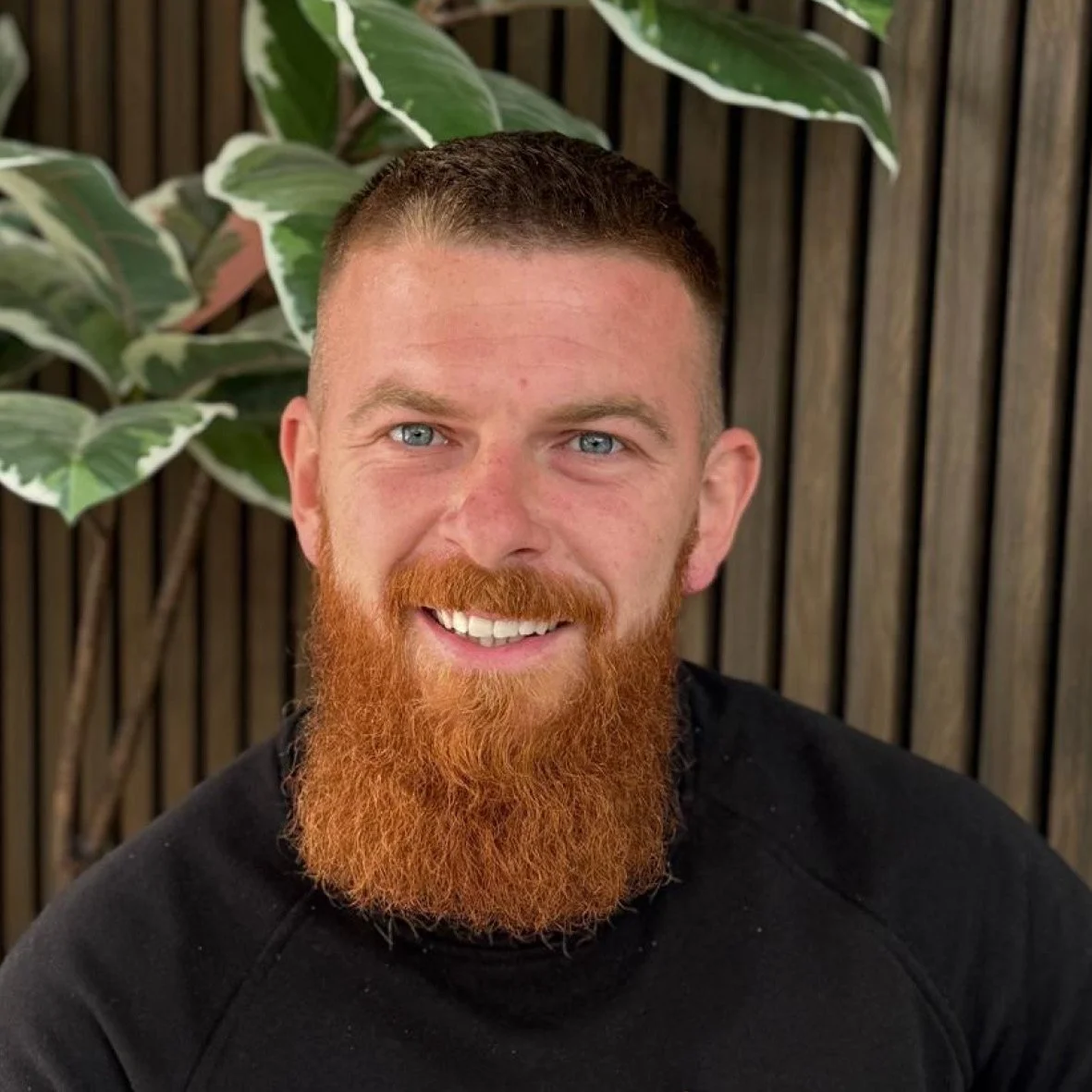 A man with a ginger beard and short brown hair smiling, seated in front of a leafy green plant and a wooden fence.