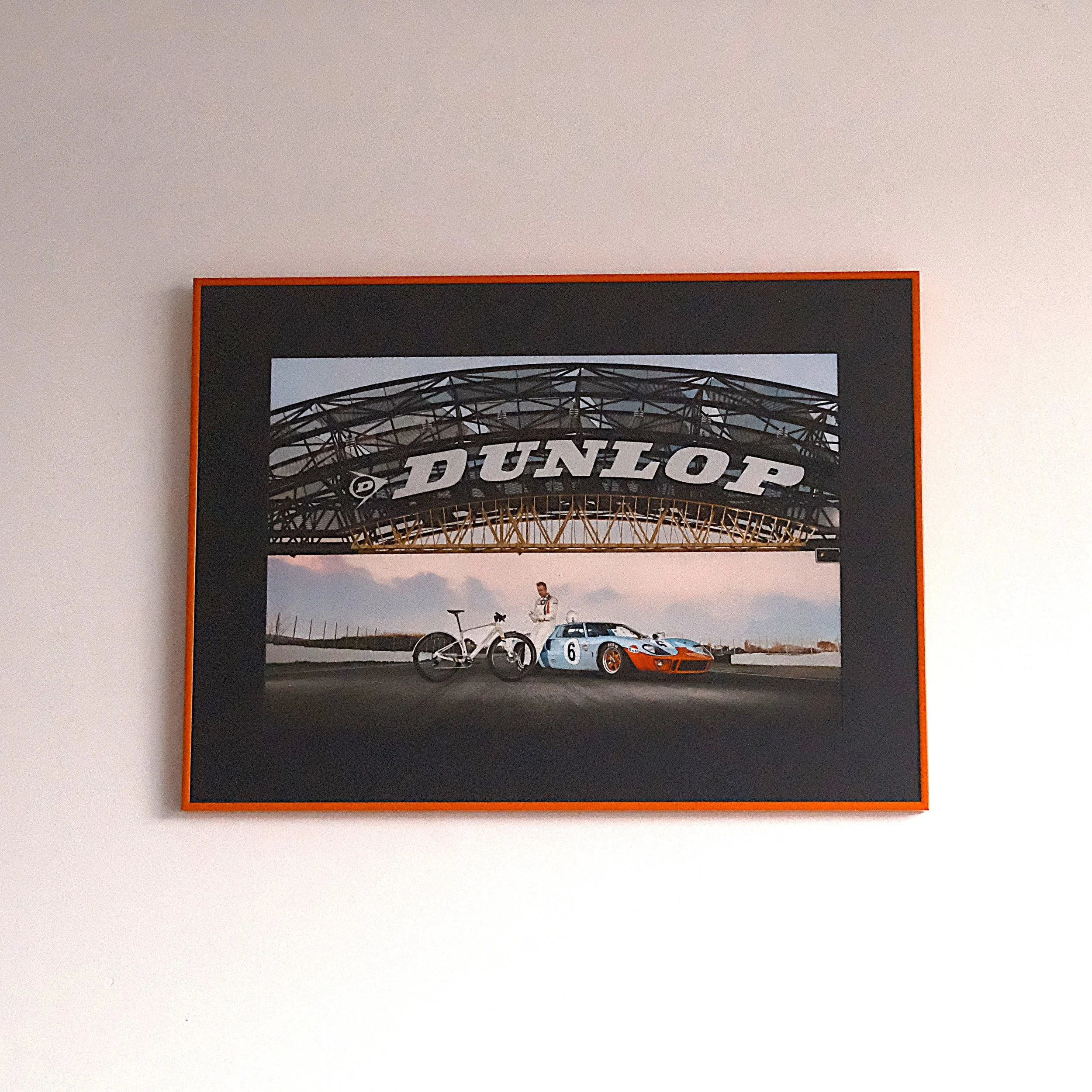 Photograph of a racing car, a cyclist, and a person standing under a large Dunlop sign at a race track during sunset.