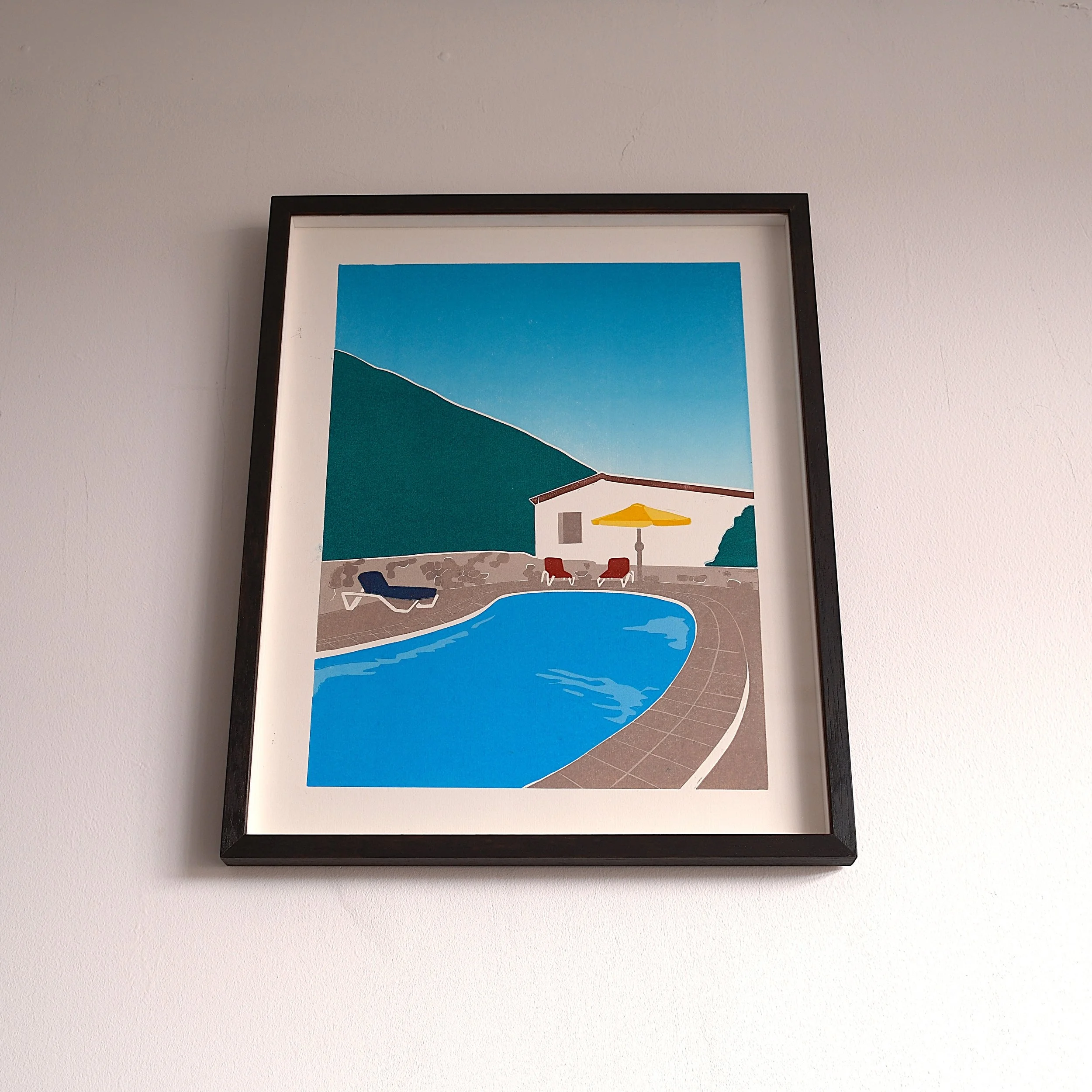 A framed illustration of a backyard swimming pool scene with a house, lounge chairs, and an umbrella under a blue sky with mountains in the background.