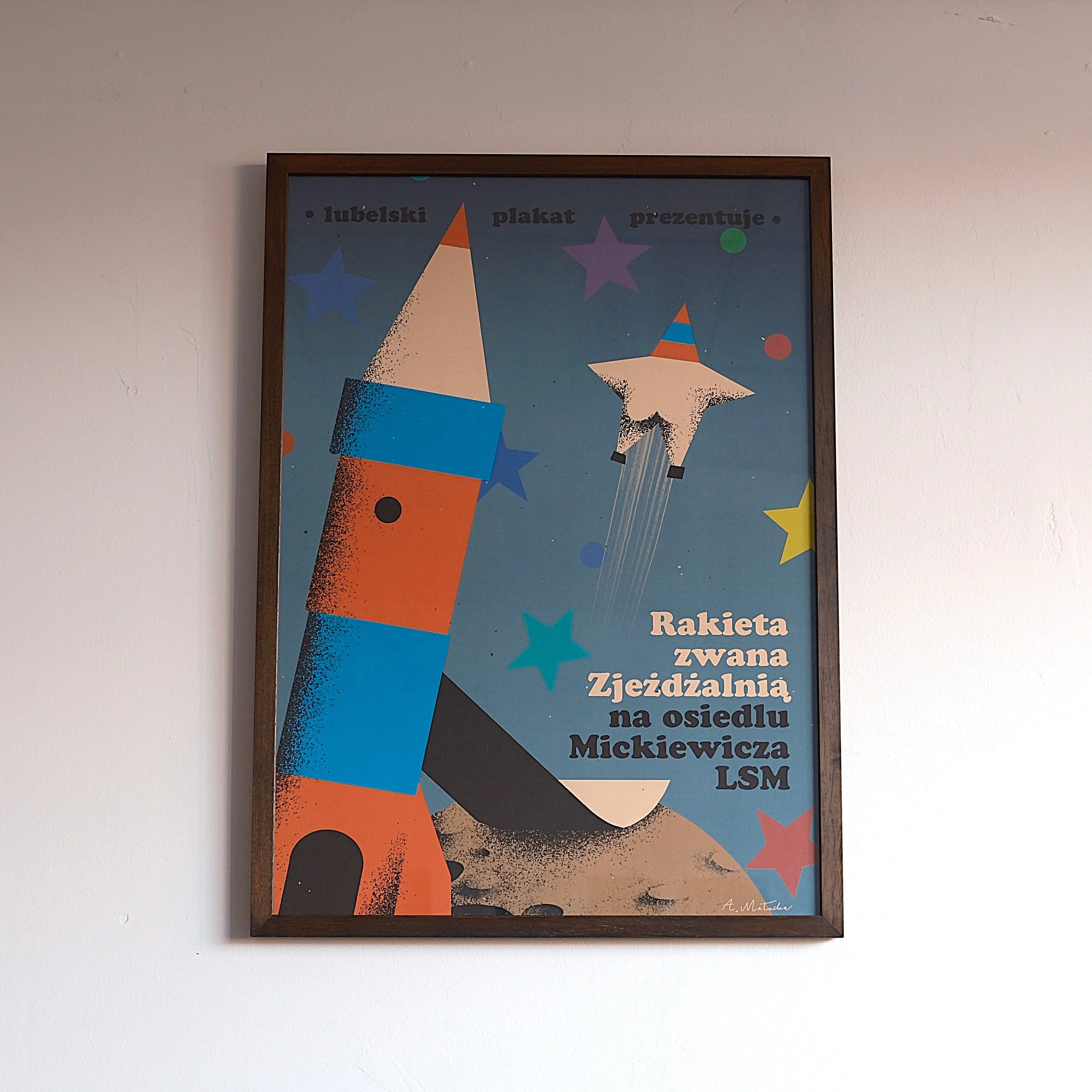 A poster with a cartoon rocket and a star-shaped rocket flying among colorful stars and dots. Text in Polish reads: "Raketa zwana Zjeżdżalnią na osiedlu Mickiewicza LSM," indicating a place named Rocket called Slide at Mickiewicz estate.