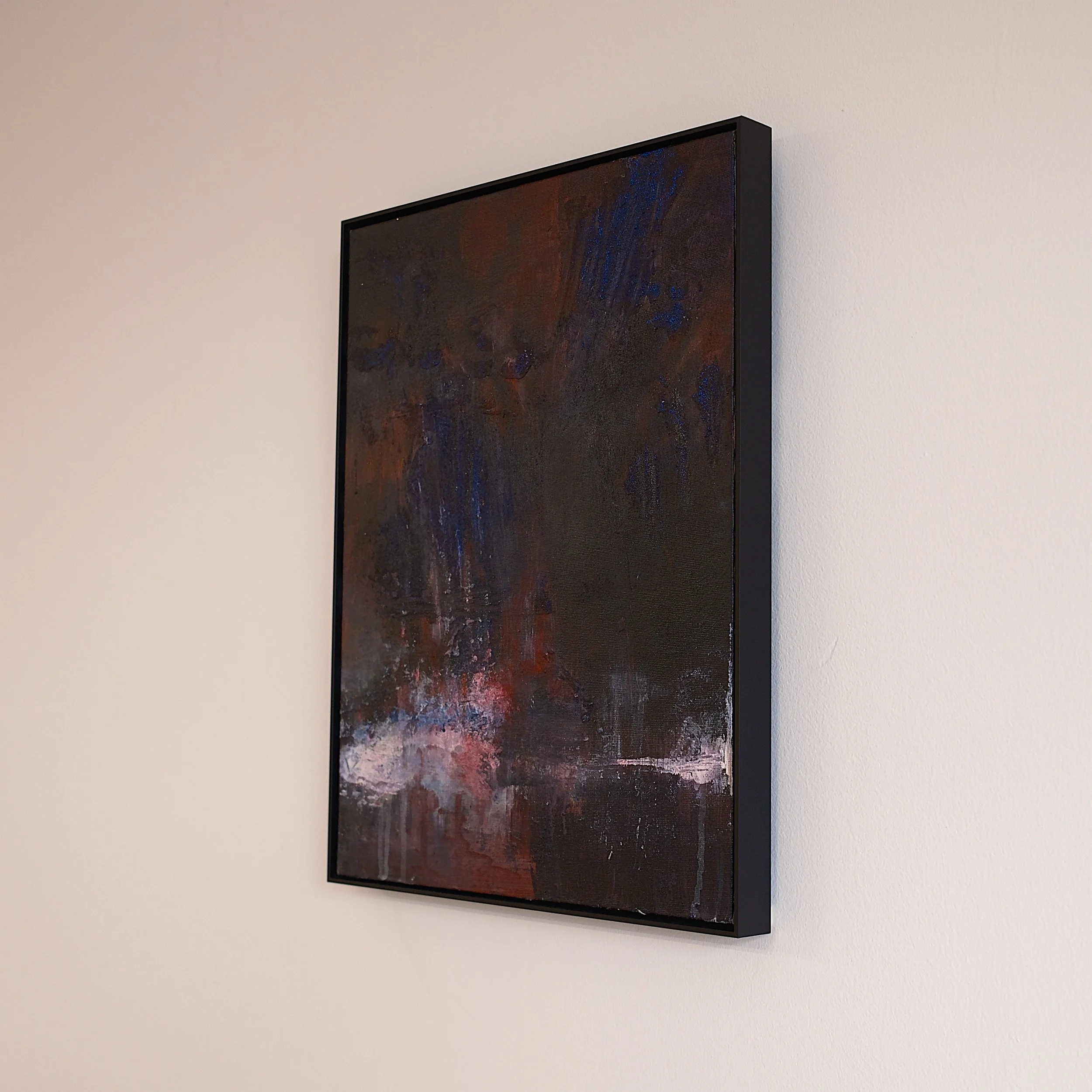 An abstract painting with dark colors, including blue, brown, black, and hints of pink and white, framed in black and hanging on a white wall.