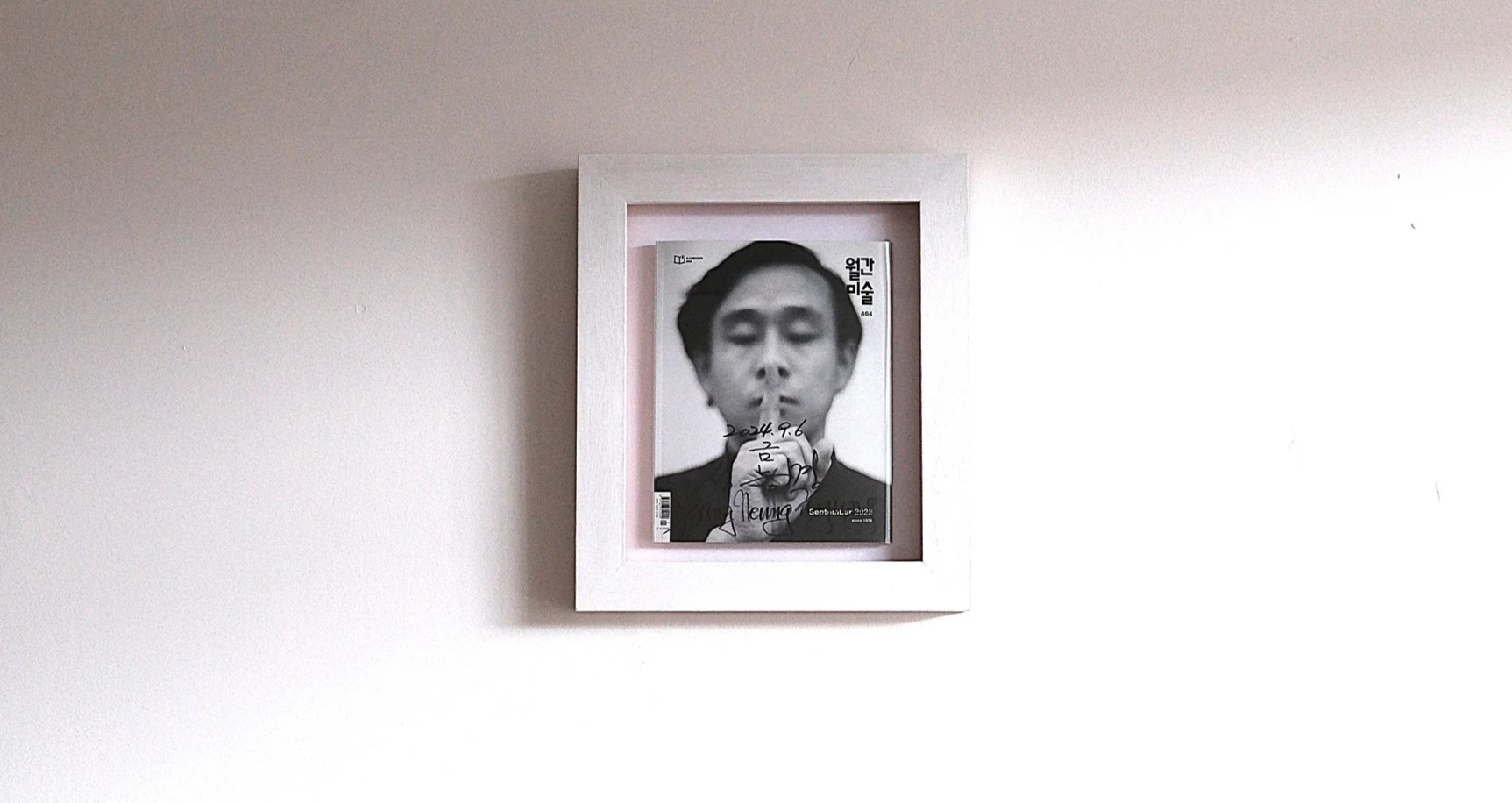 A framed magazine cover on a white wall featuring a black-and-white portrait of a woman with short hair, eyes closed, and a finger to her lips, with handwritten text and signatures on the cover.