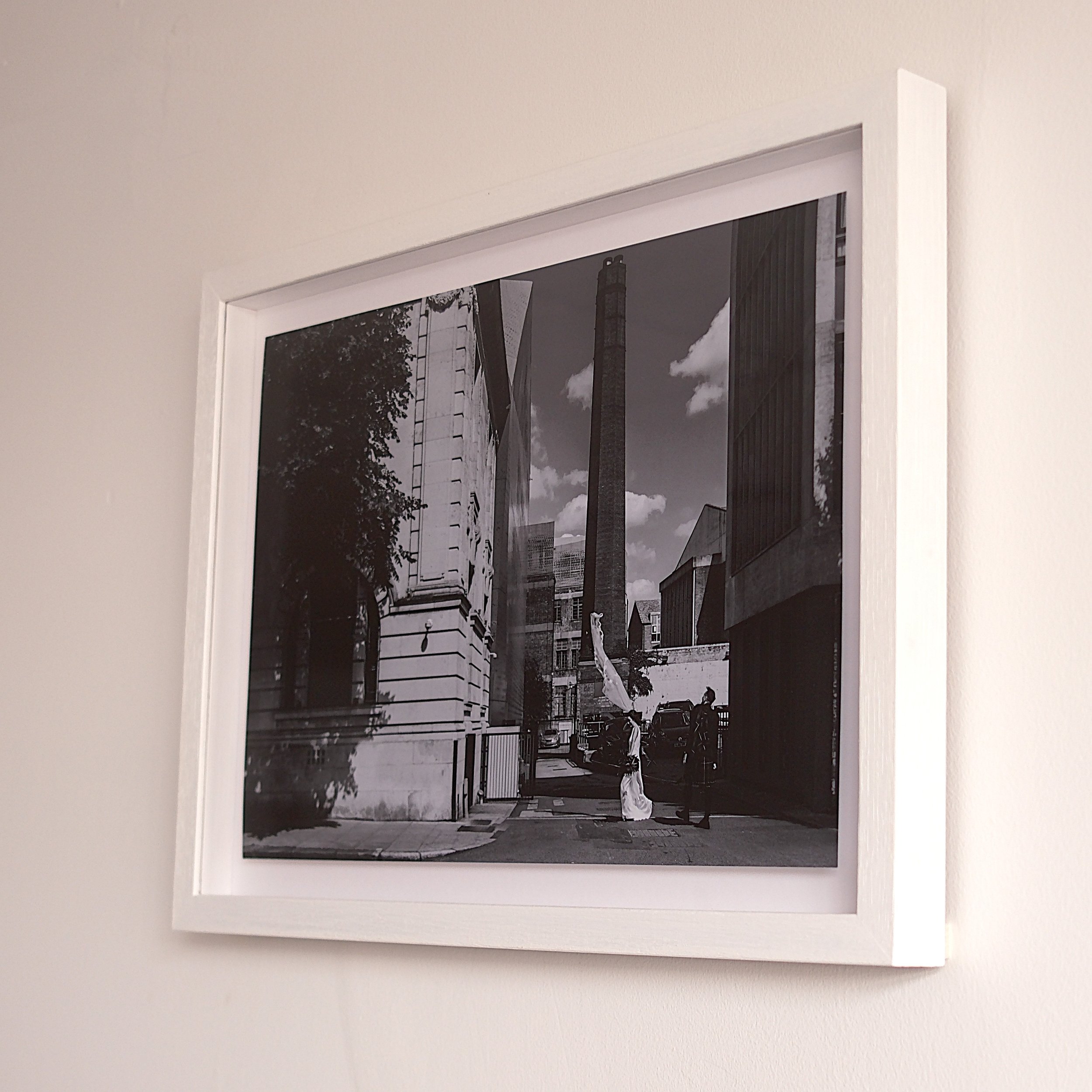 Black and white photograph of an urban street scene with tall buildings, a chimney, and two people walking, framed and hanging on a white wall.
