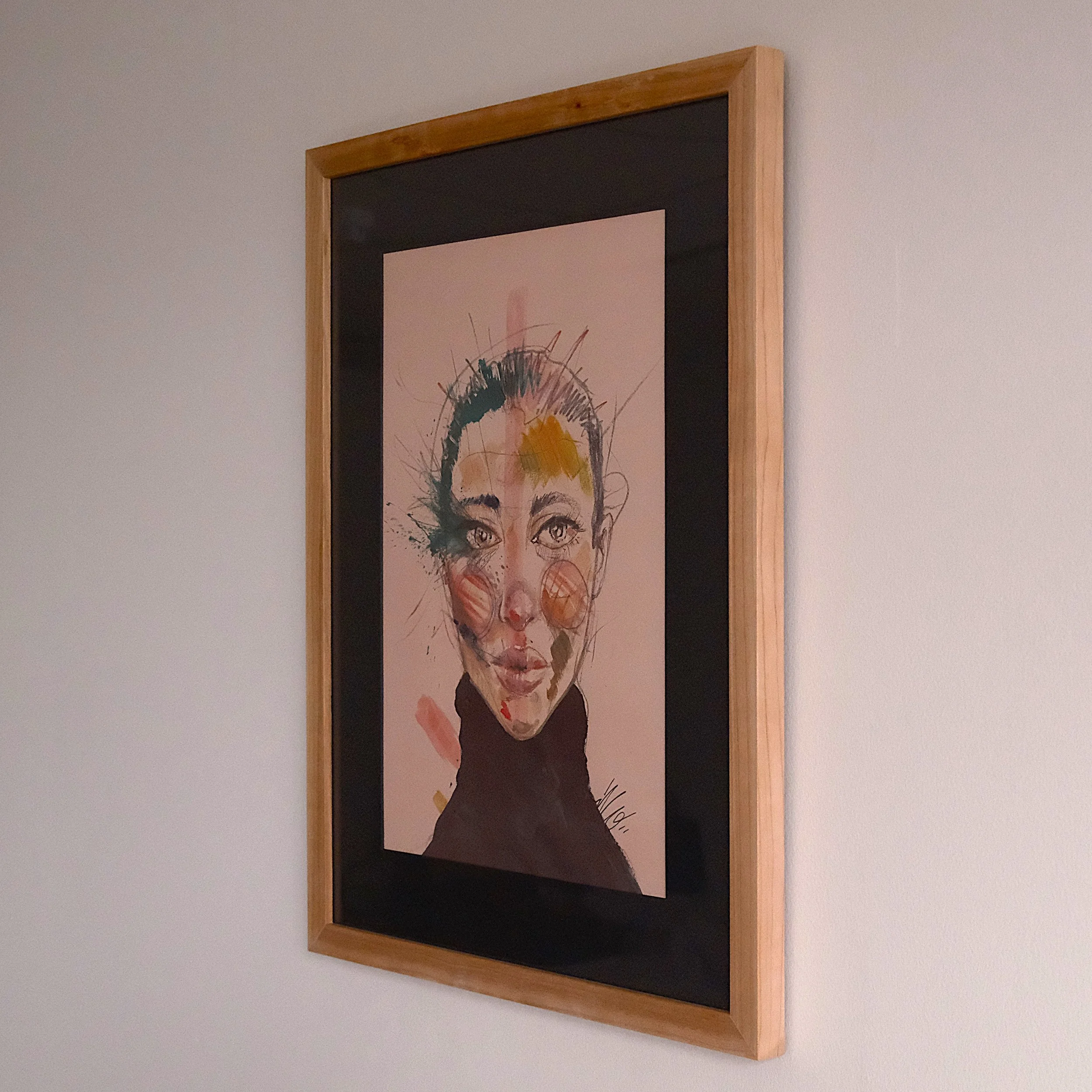 Watercolor portrait of a woman with short hair, wearing a black turtleneck, with colorful abstract splashes on her face, framed in a wooden frame hanging on a white wall.
