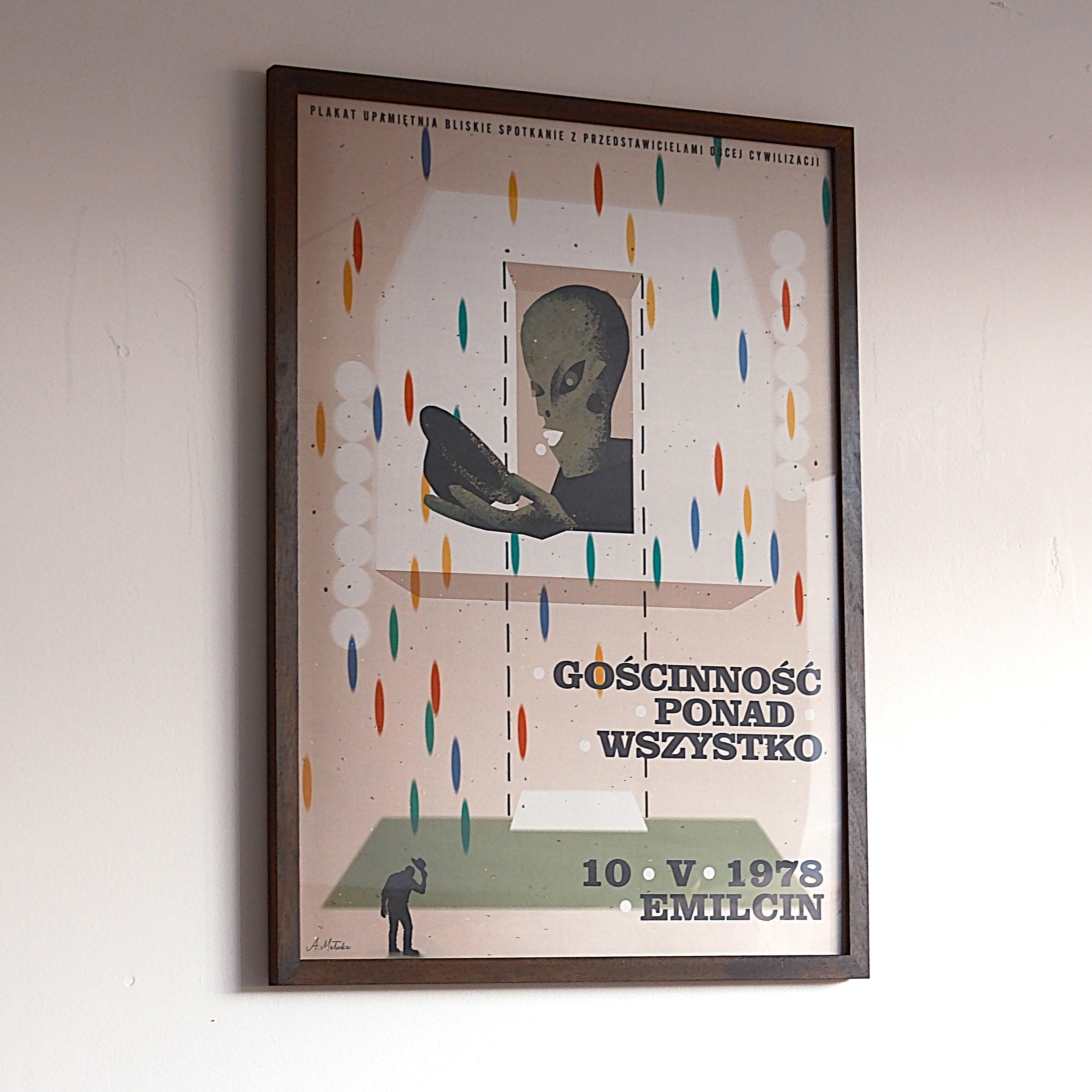 Poster featuring an alien reading a book, with colorful abstract shapes around, and a small silhouette of a person at the bottom left. Text in Polish: "Gościnność ponad wszystko," with a date "10 V 1978" and location "Emilcin."