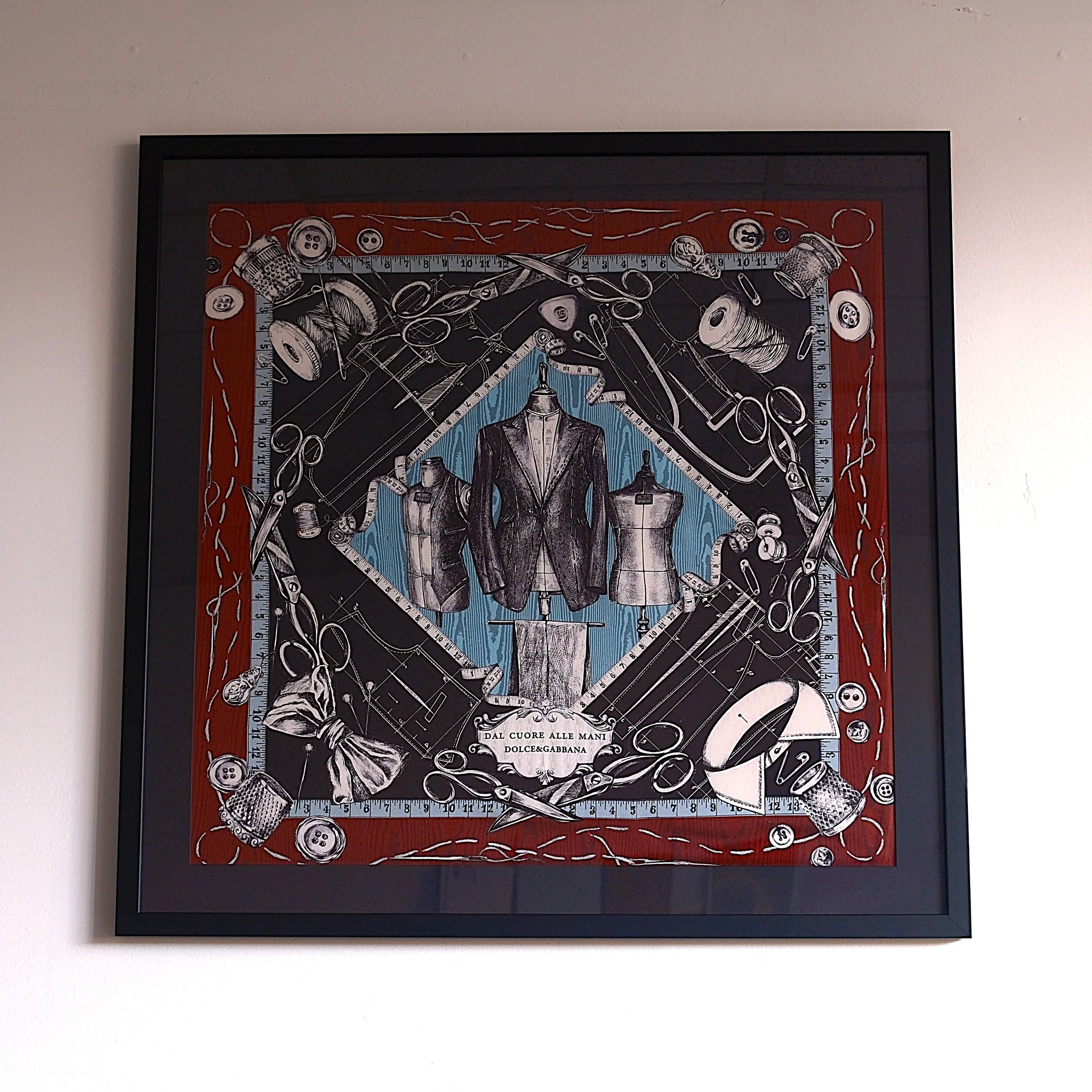 A framed artwork featuring sewing tools, fabric swatches, and mannequins, with a central fashion suit display on a mannequin, and Italian text at the bottom.