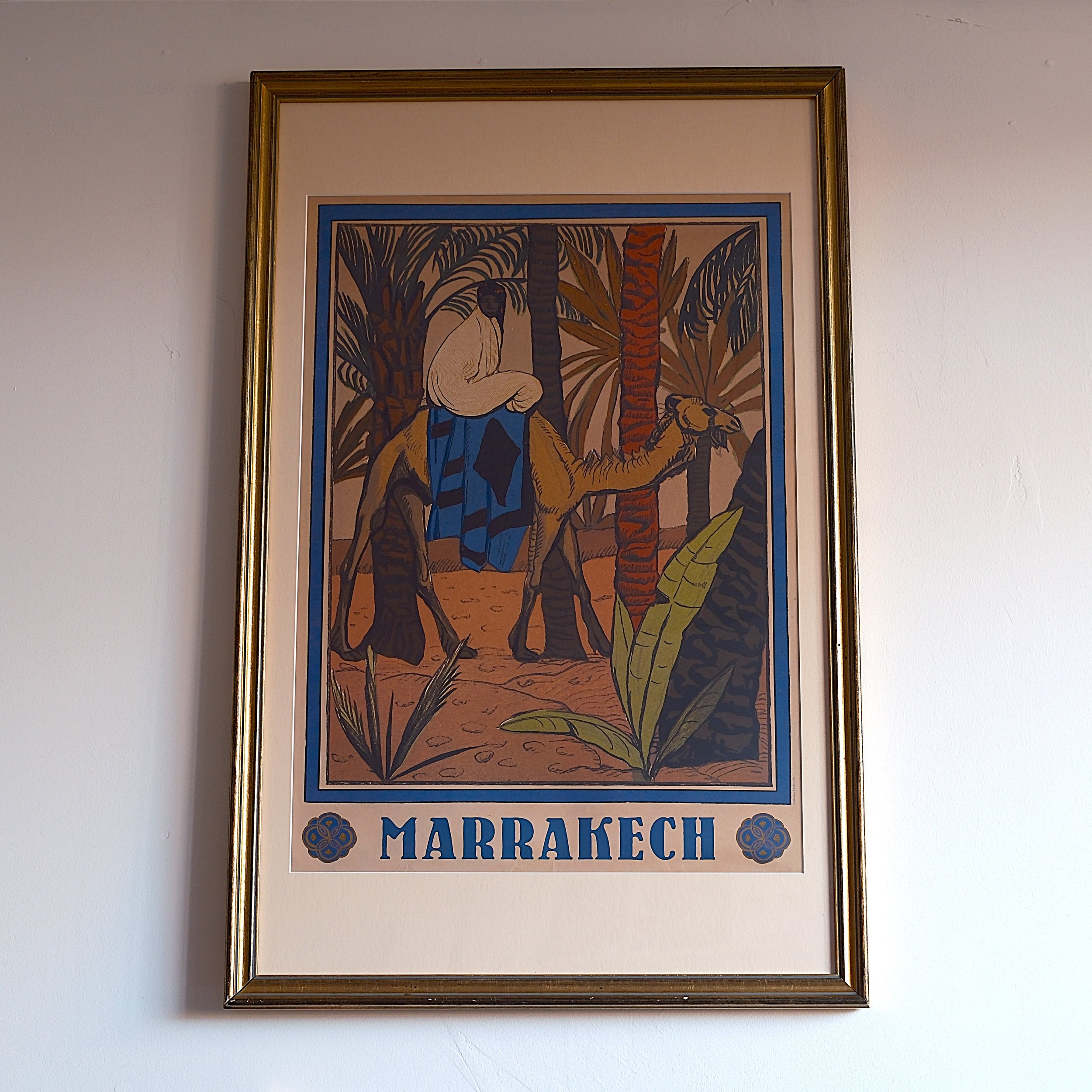 A framed vintage poster of Marrakech featuring a person riding a camel through a desert with palm trees and lush vegetation in the background.