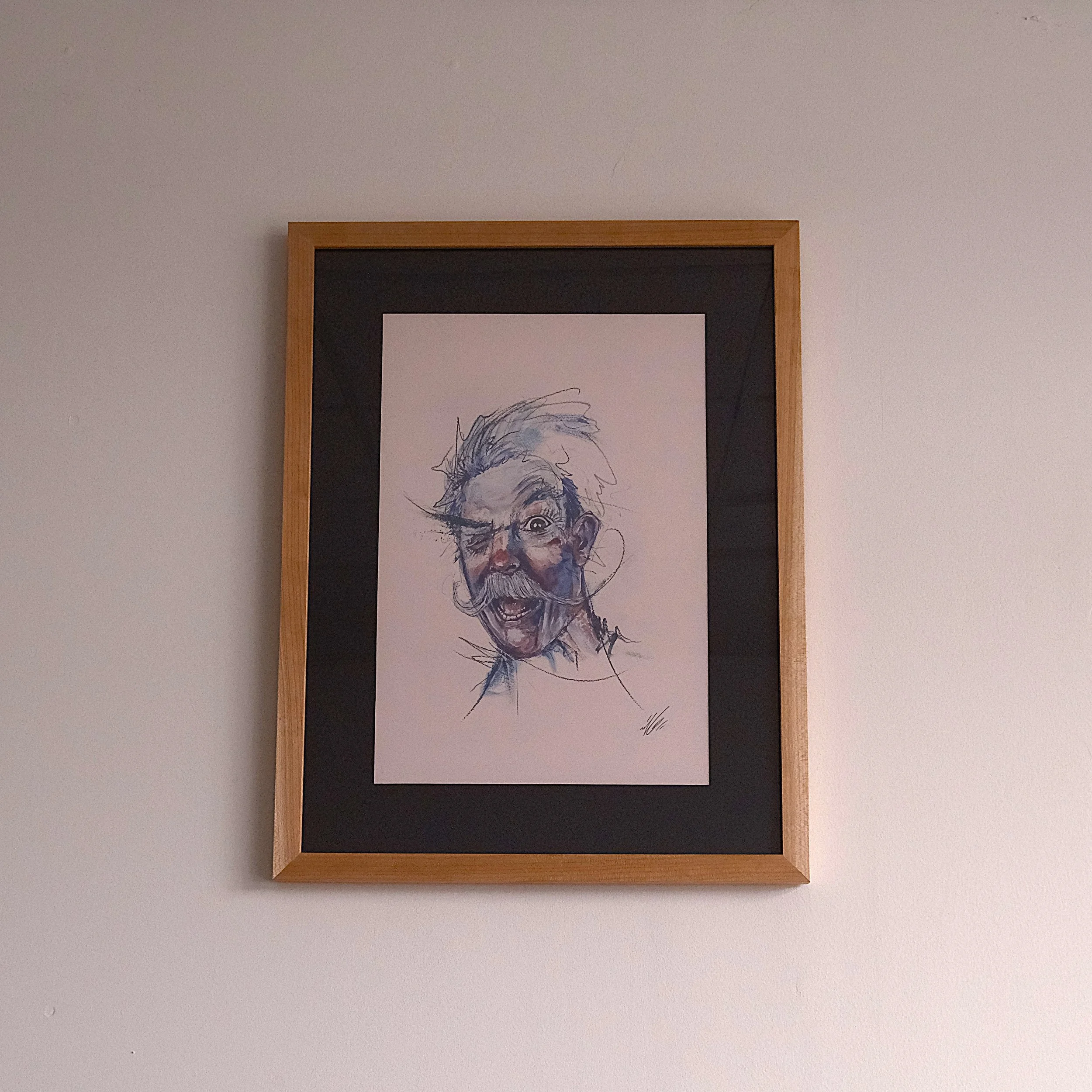 A framed, colorful caricature drawing of a man with wild hair, a mustache, and beard on a white wall.
