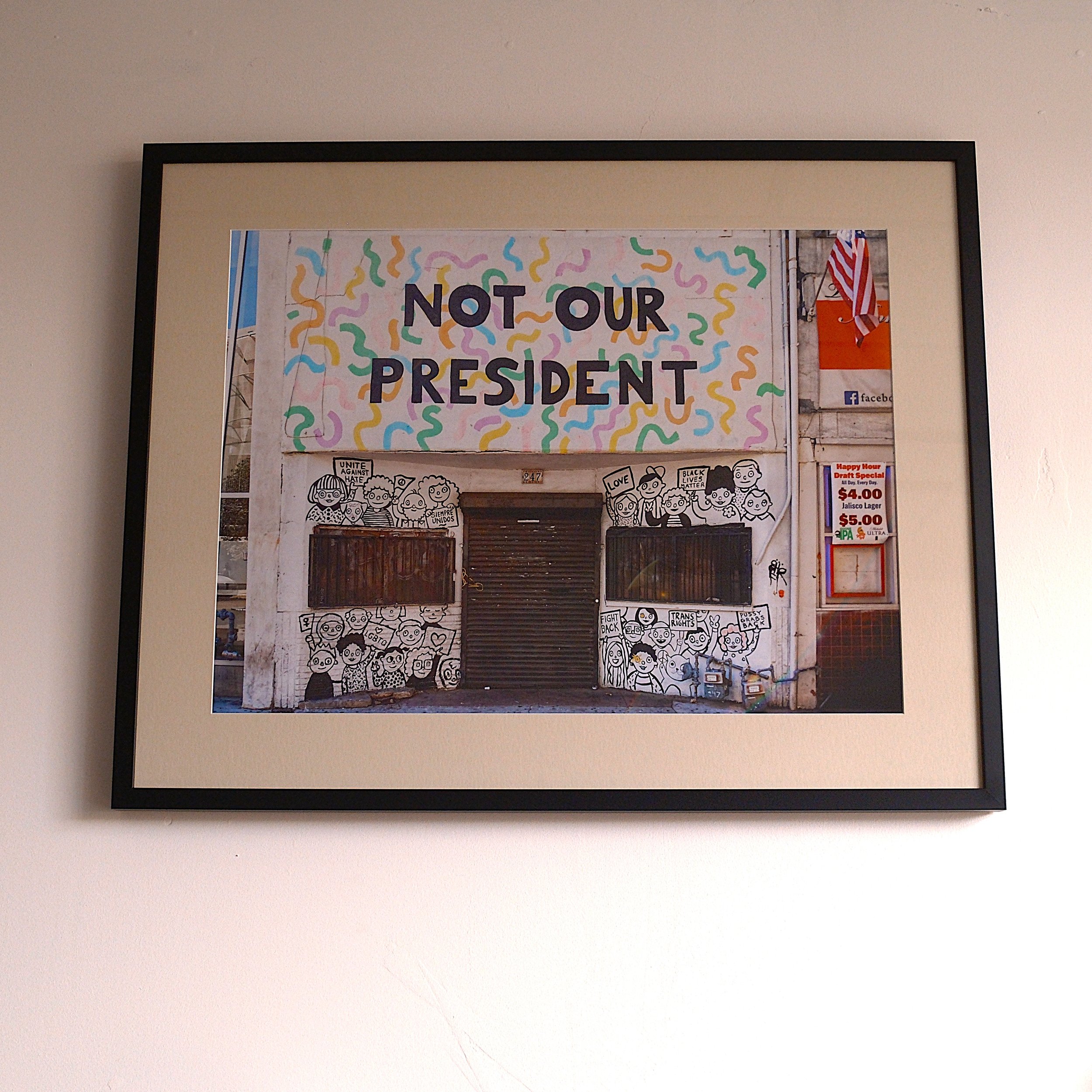 A framed photo of a storefront with a large sign that reads 'NOT OUR PRESIDENT'. The storefront has cartoon-style illustrations of diverse people protesting, holding signs with messages like 'LOVE', 'UNITE', and 'FIGHT BACK'. Nearby are signs advertising prices for beverages.