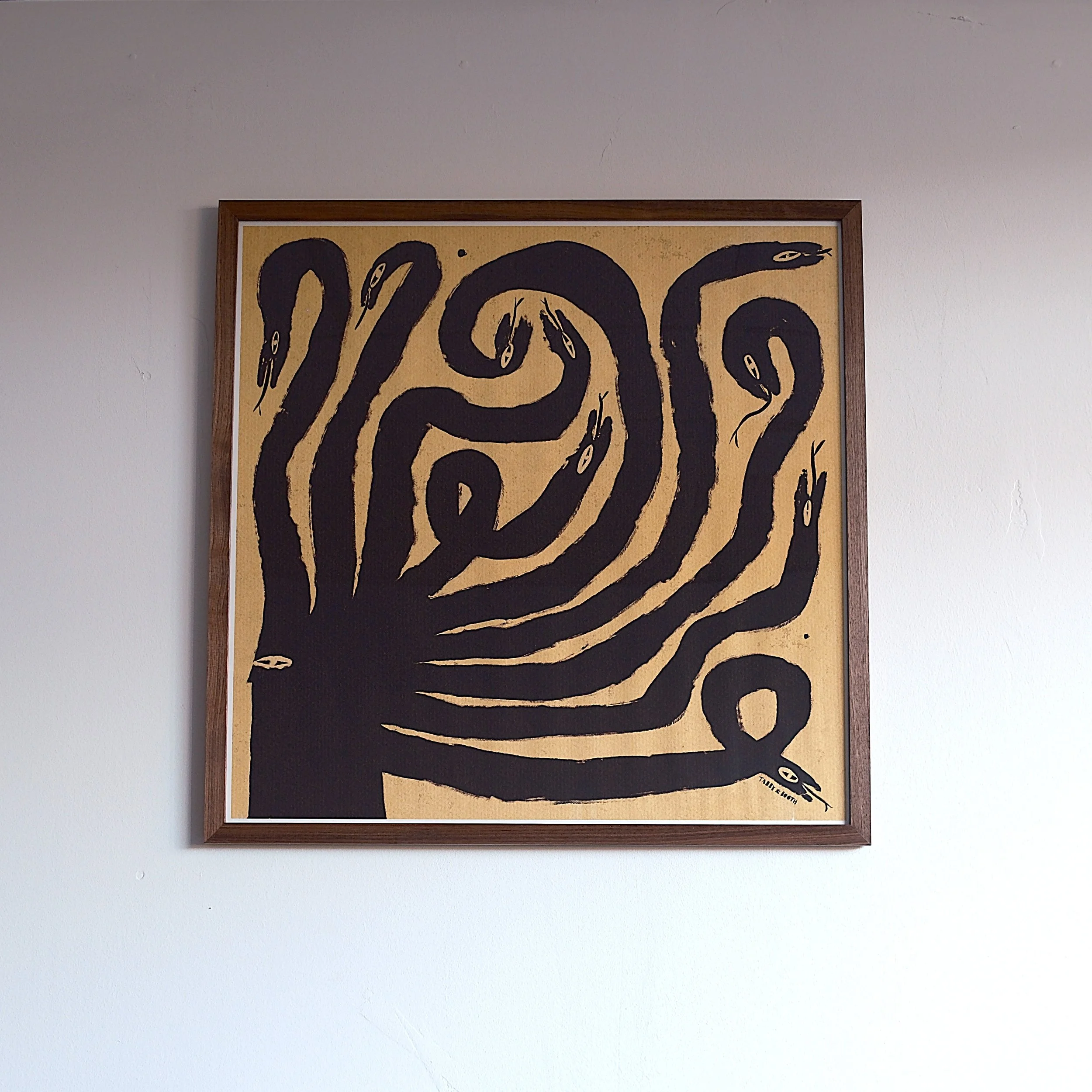 A framed artwork of a black snake with multiple heads on a tan background, mounted on a white wall.