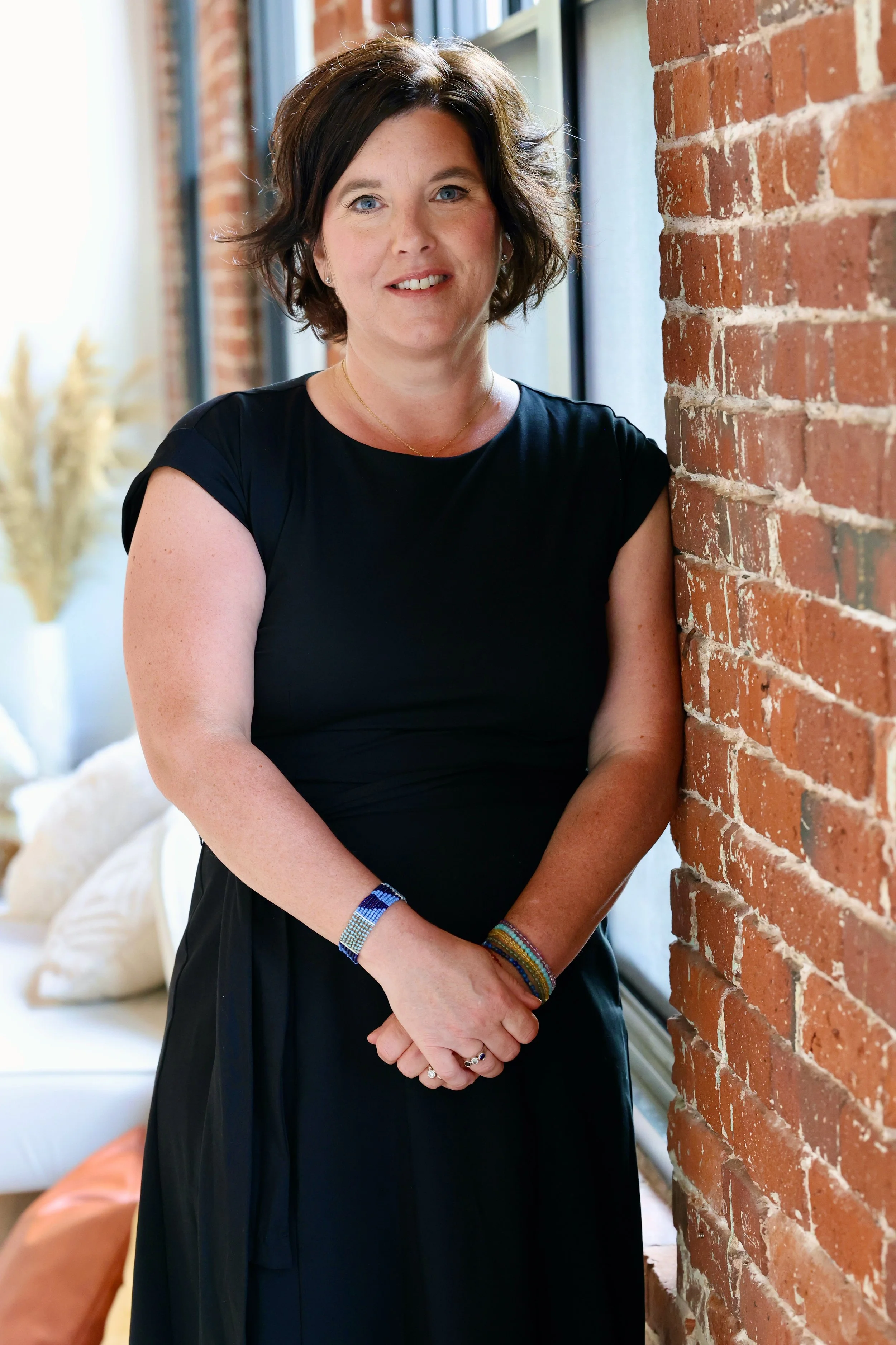 Angela Gowans with short brown hair, blue eyes, and light skin, wearing a black dress, standing indoors next to a brick wall, smiling at the camera.
